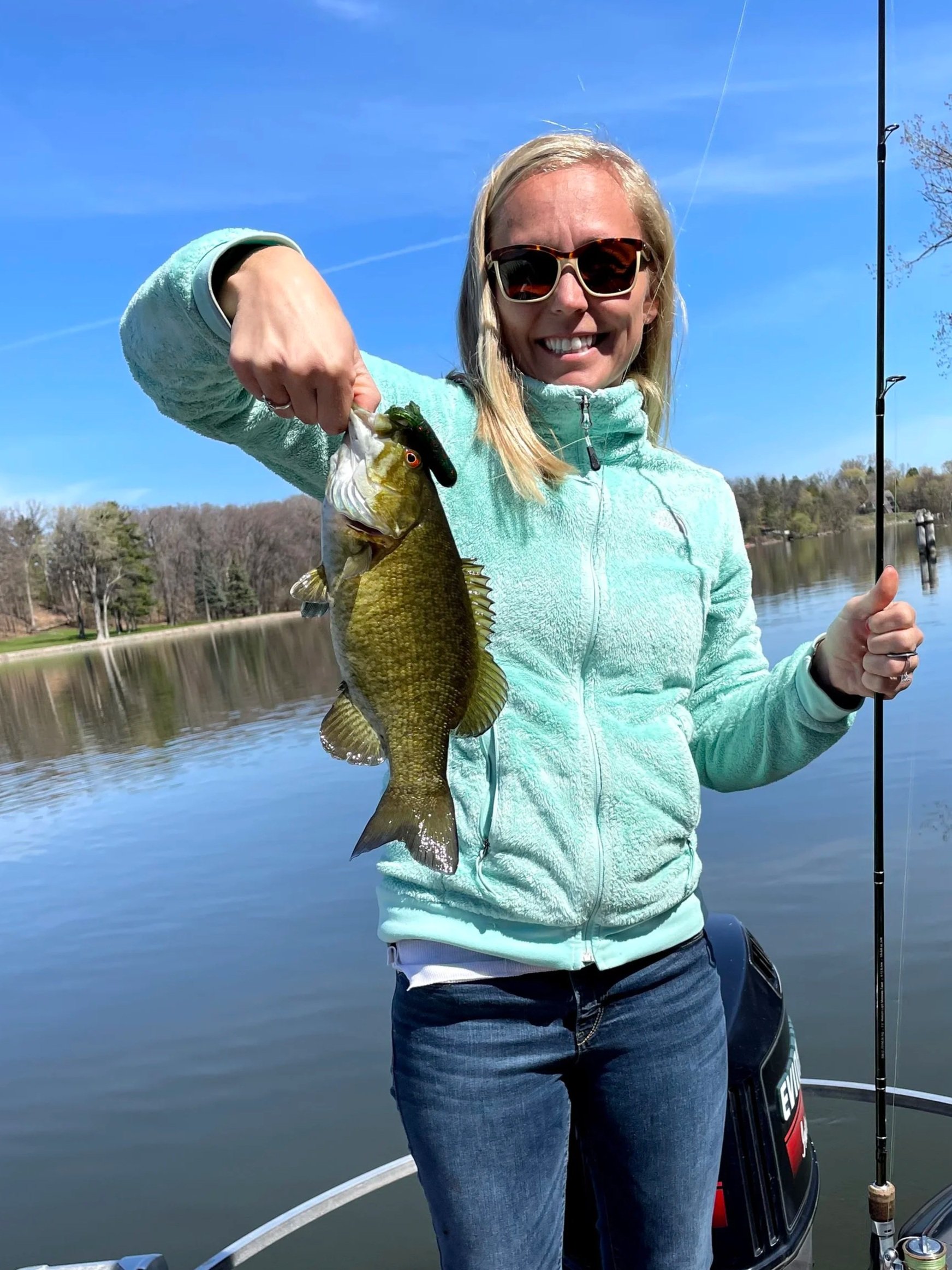 Woman smiling, wearing sunglasses and a light blue jacket, holding a fish she caught on a fishing trip by a lake.