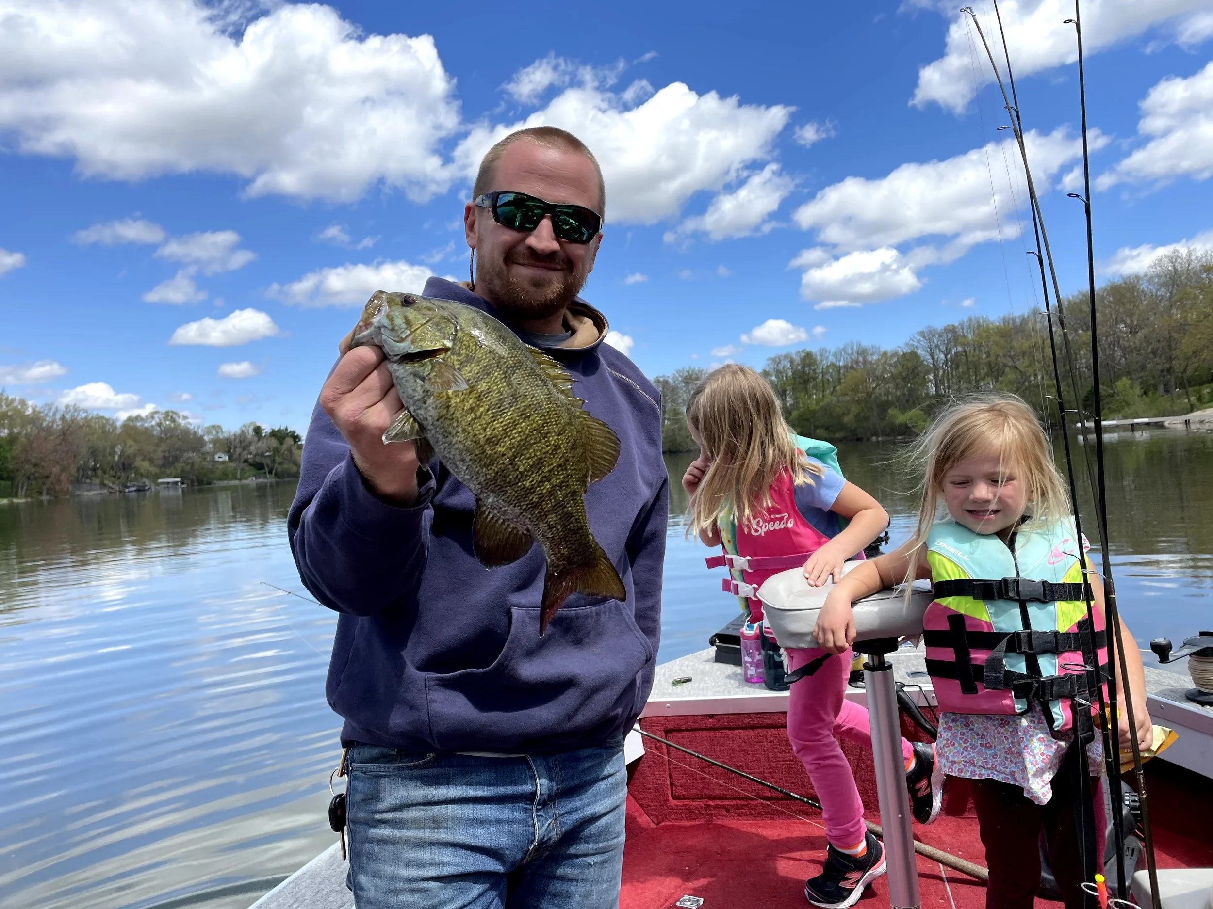A man with sunglasses holding a fish on a boat with two young girls in life jackets fishing, lake and blue sky with clouds in the background.