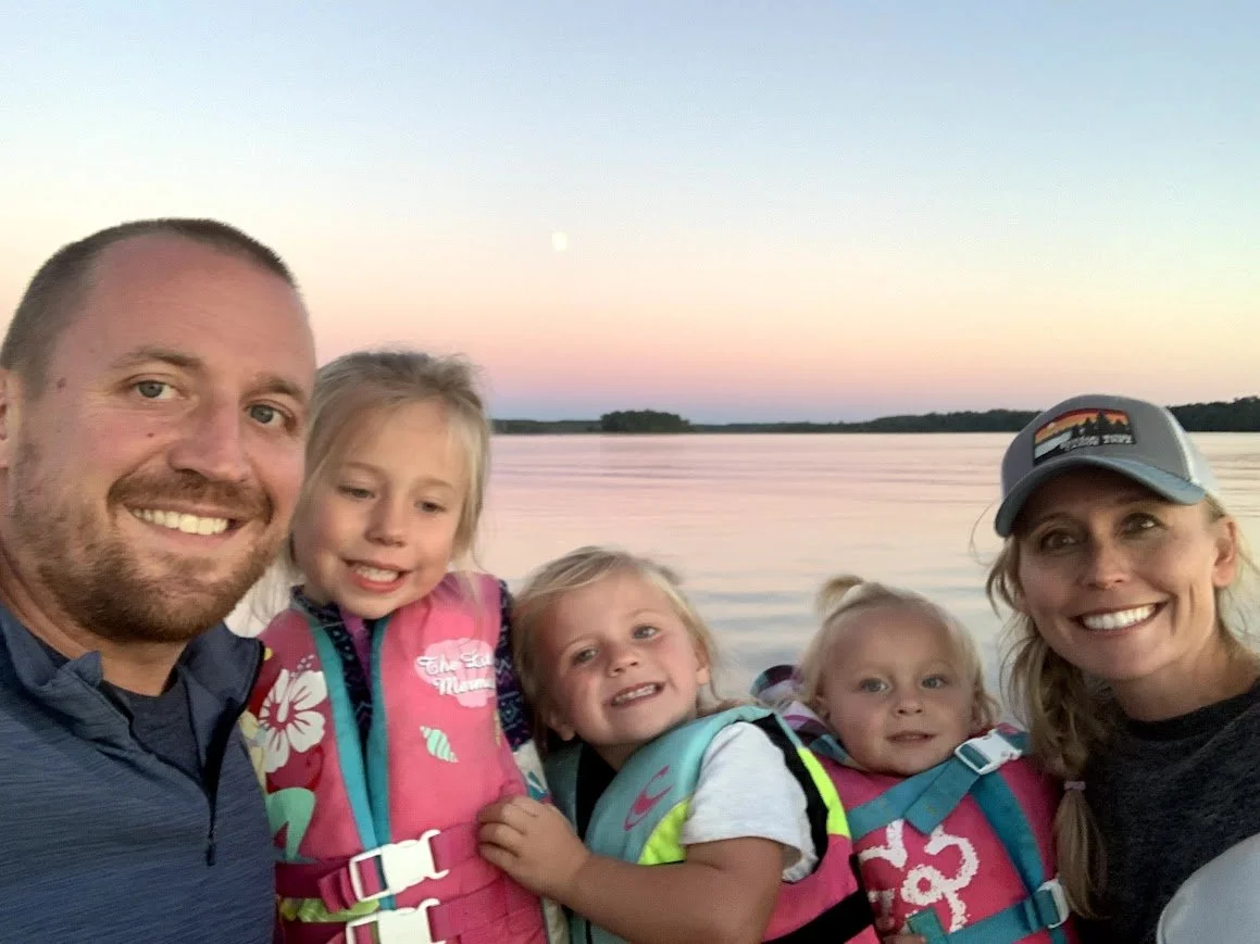 A family of five taking a selfie by a lake during sunset at the BWCA, with the moon visible in the sky. The family members include a man, a woman, and three young girls all smiling.