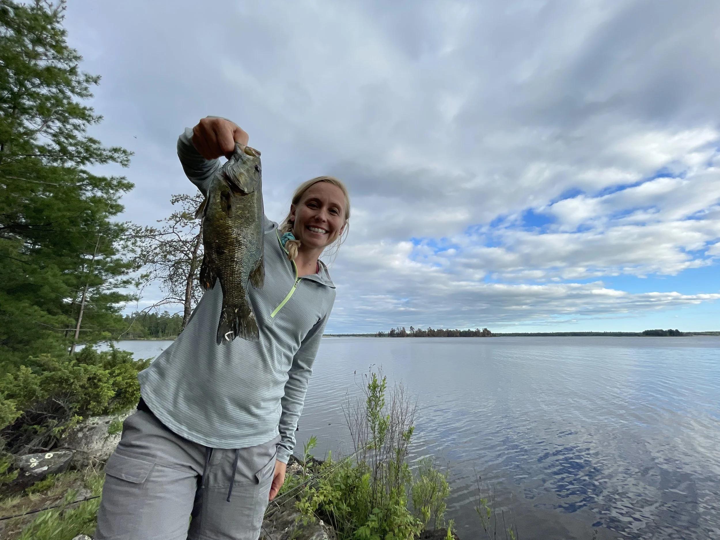 A woman holding a fishing catch of a largemouth bass by a lake on a partly cloudy day.