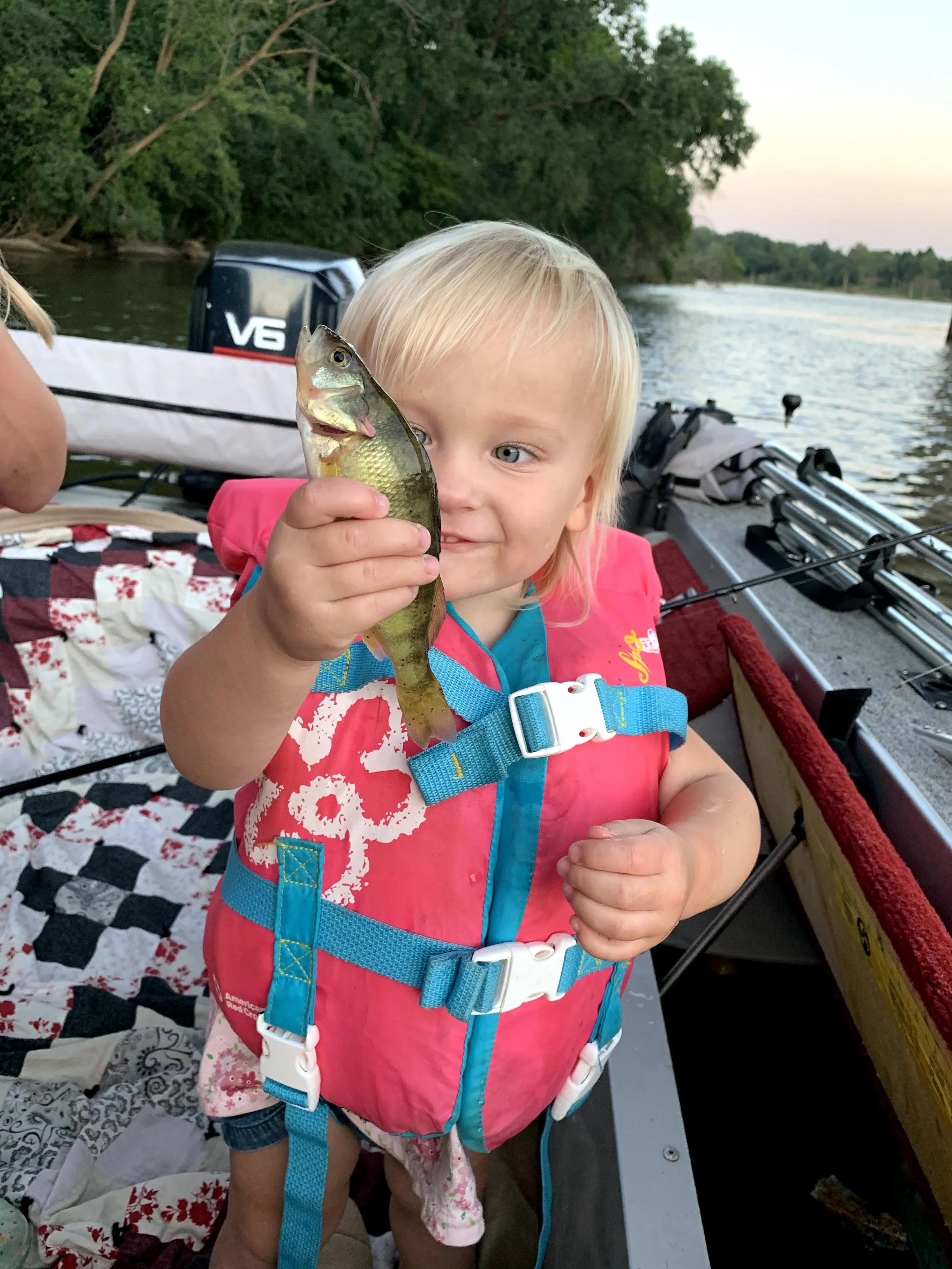 A young girl in a pink life jacket holding a perch on a boat with trees and water in the background.