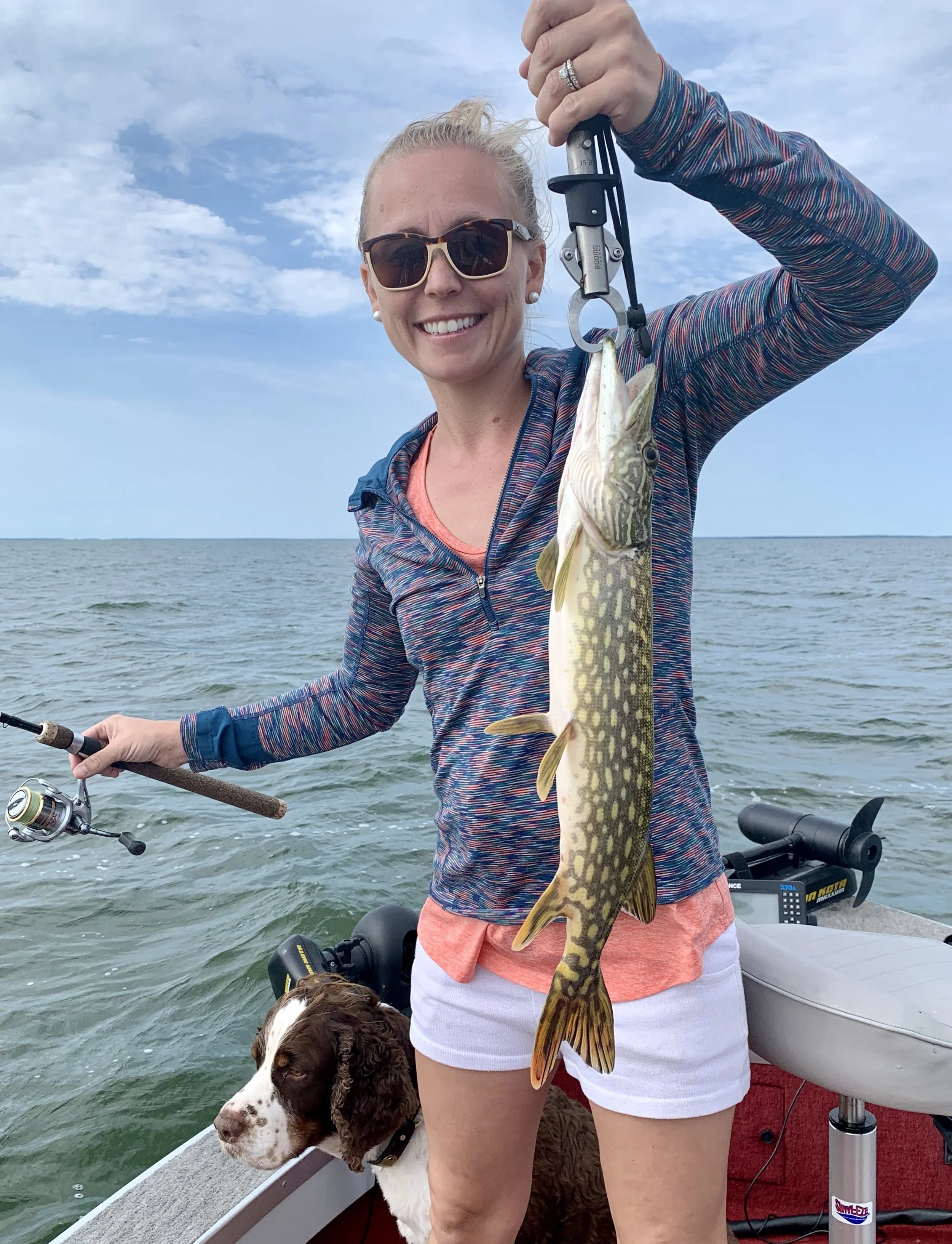 A woman with sunglasses holding a large Northern Pike on a boat, with a dog sitting nearby, in an open water setting under cloudy skies.