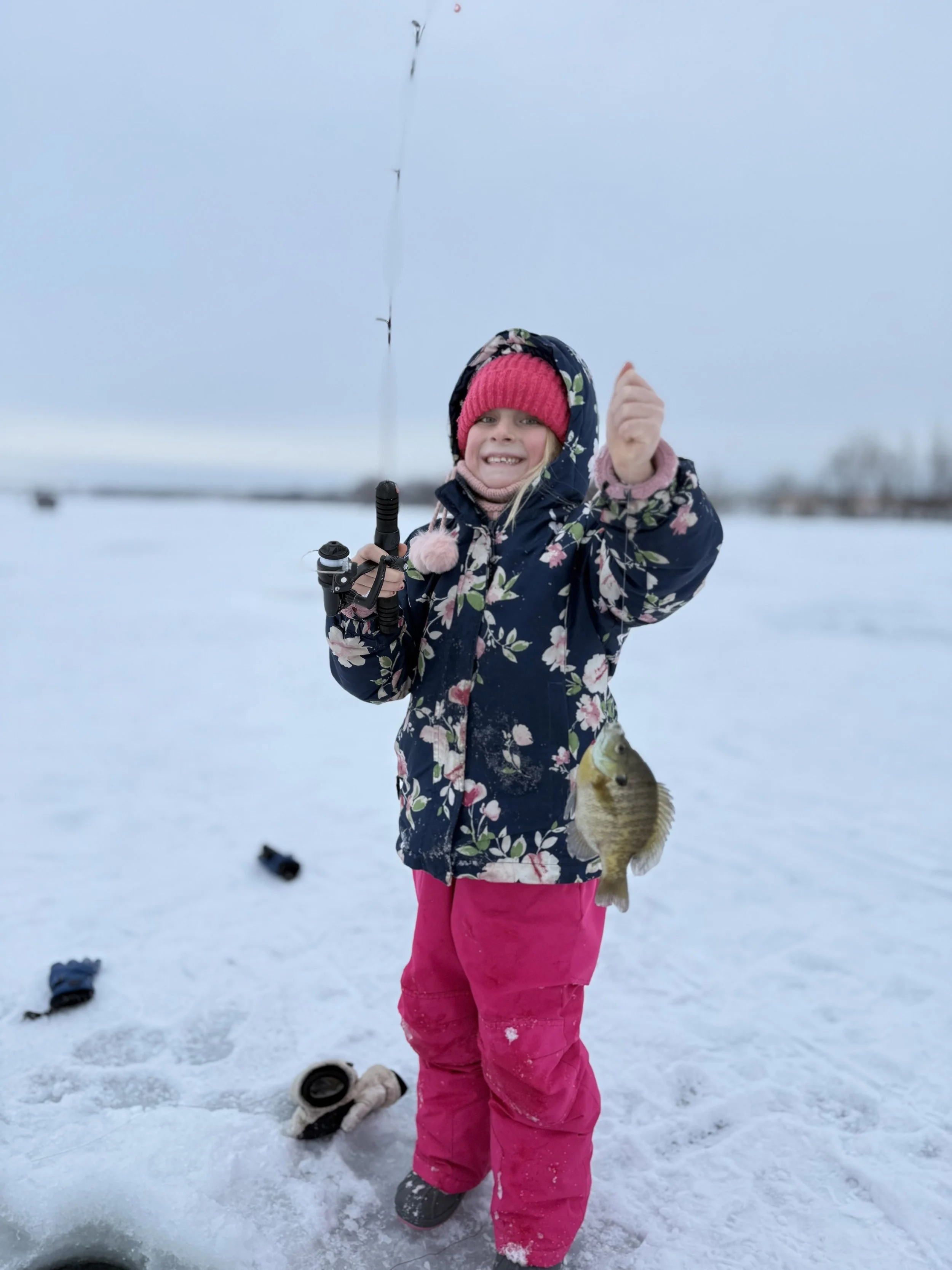 Girl in winter clothing holding a fishing rod with a fish, standing on snow-covered ice outdoors.