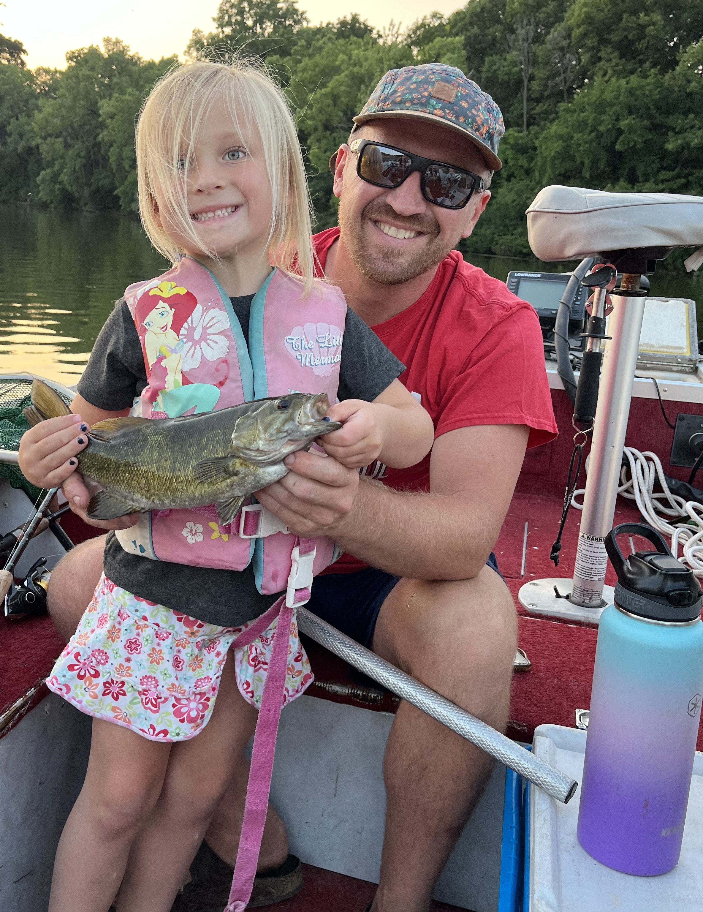 A young girl and a man on a boat holding a fish, with trees and water in the background.