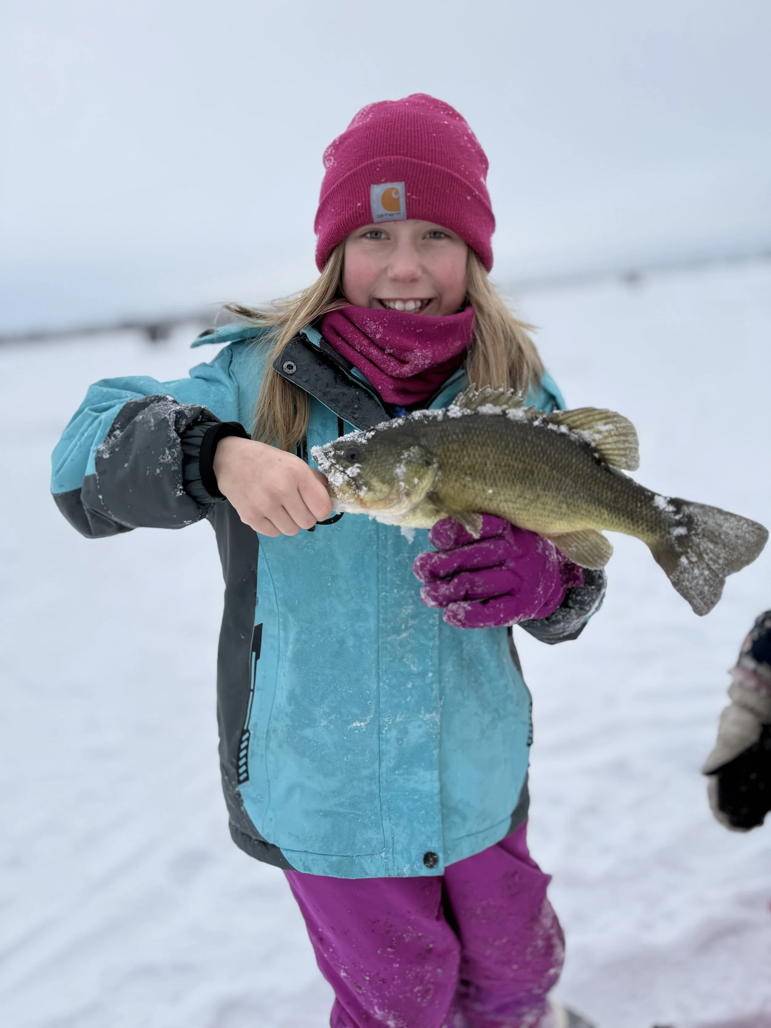 A young girl in winter clothing smiling and holding a fish she caught, standing in a snowy landscape.