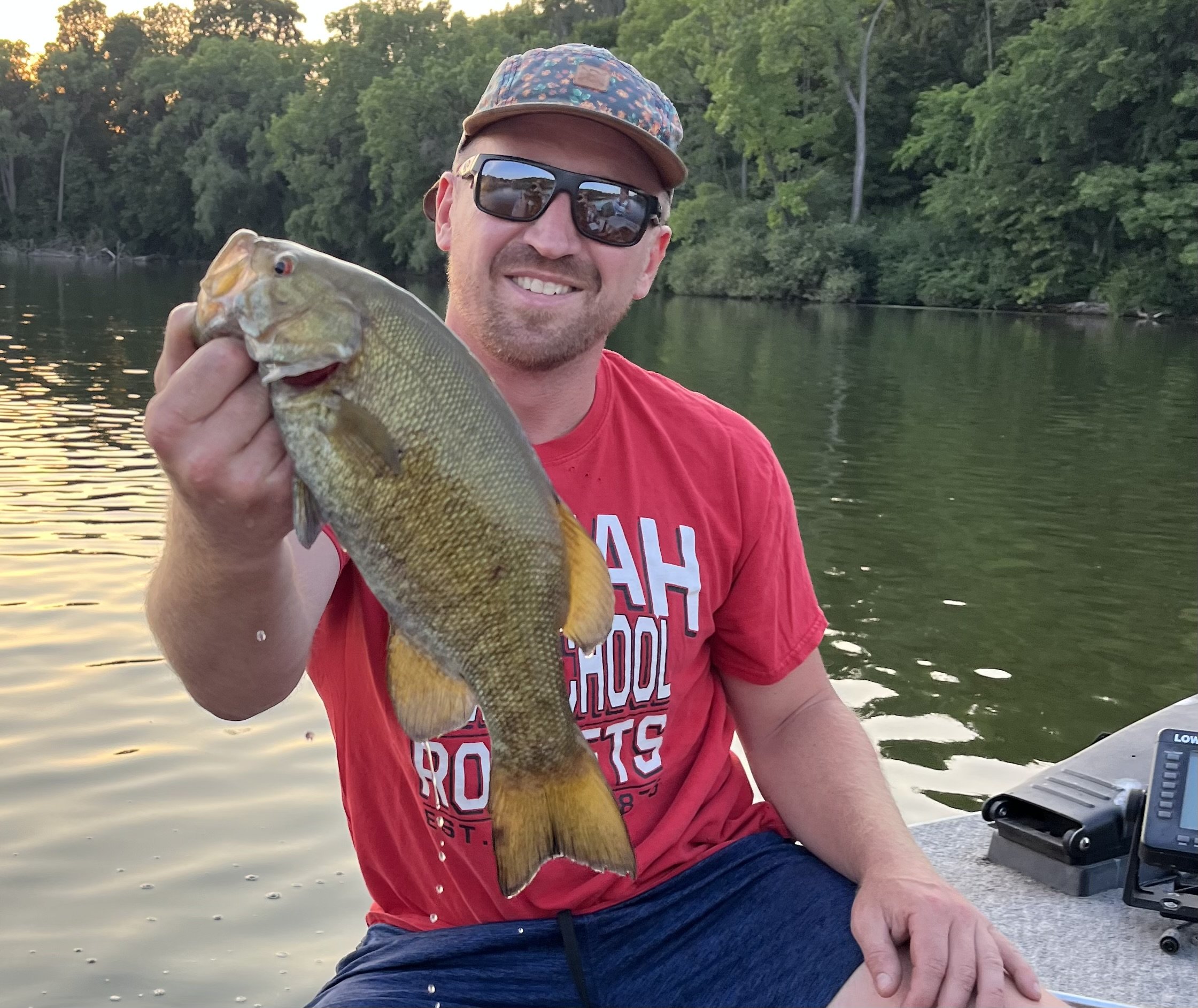 Man holding a large fish on a boat in a river, with trees and sunset in the background.