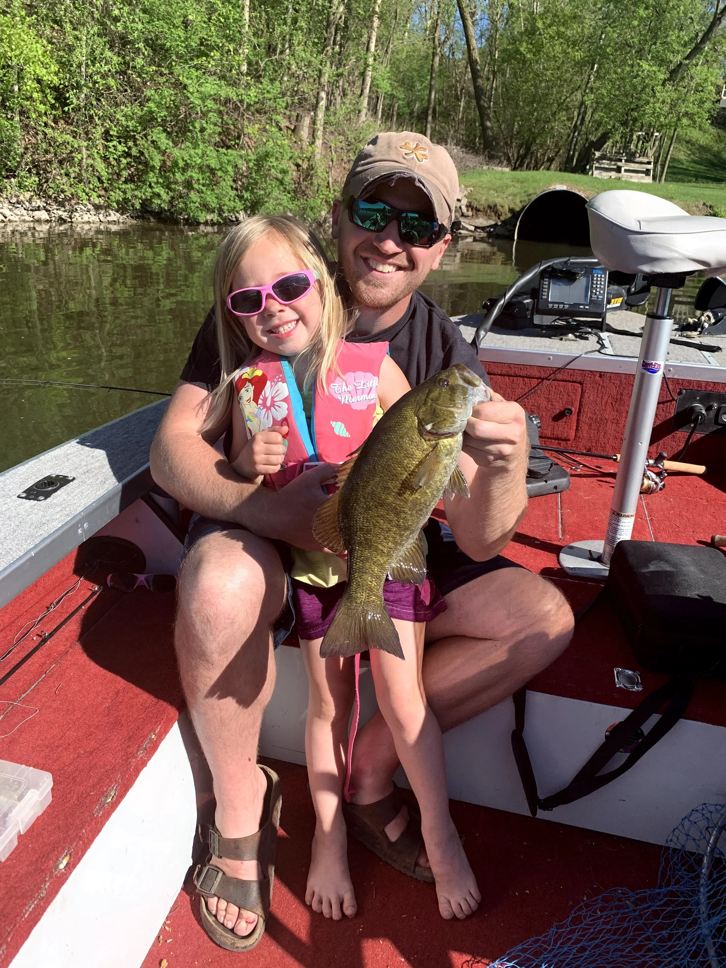 A man and a young girl sitting on a boat, holding a large fish they caught. The man is wearing a tan hat, sunglasses, and a black T-shirt, smiling at the camera. The girl is wearing pink sunglasses and a pink life jacket with a mermaid design, also s