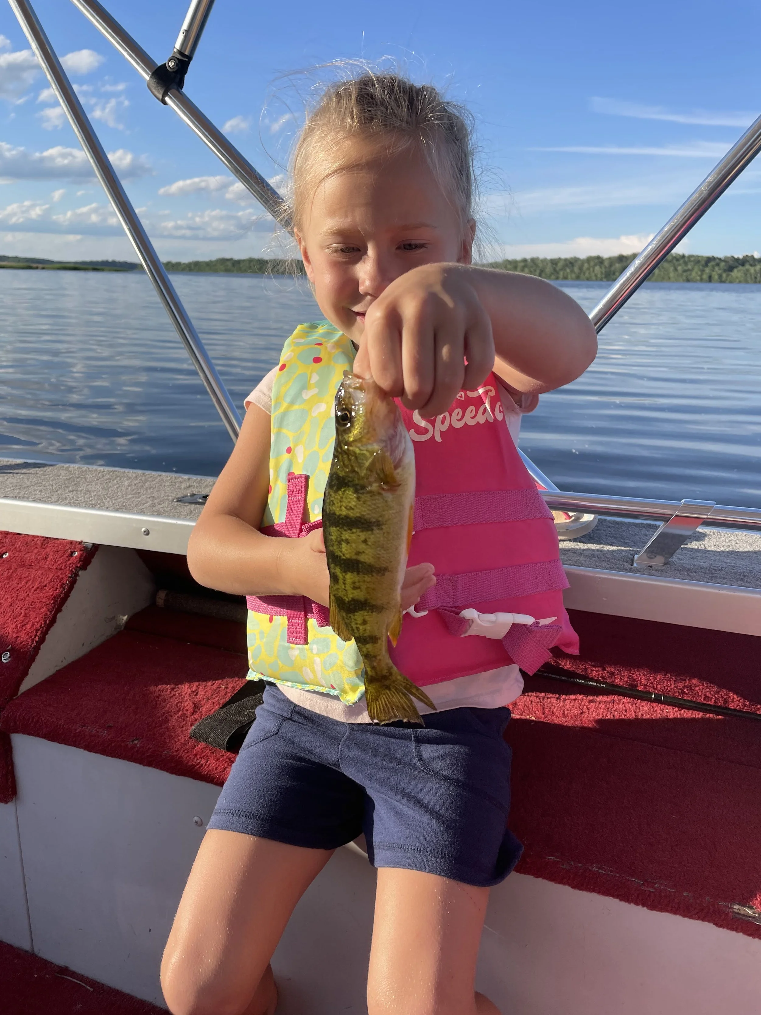 Young girl on a boat holding a perch she caught, with water and blue sky in the background.