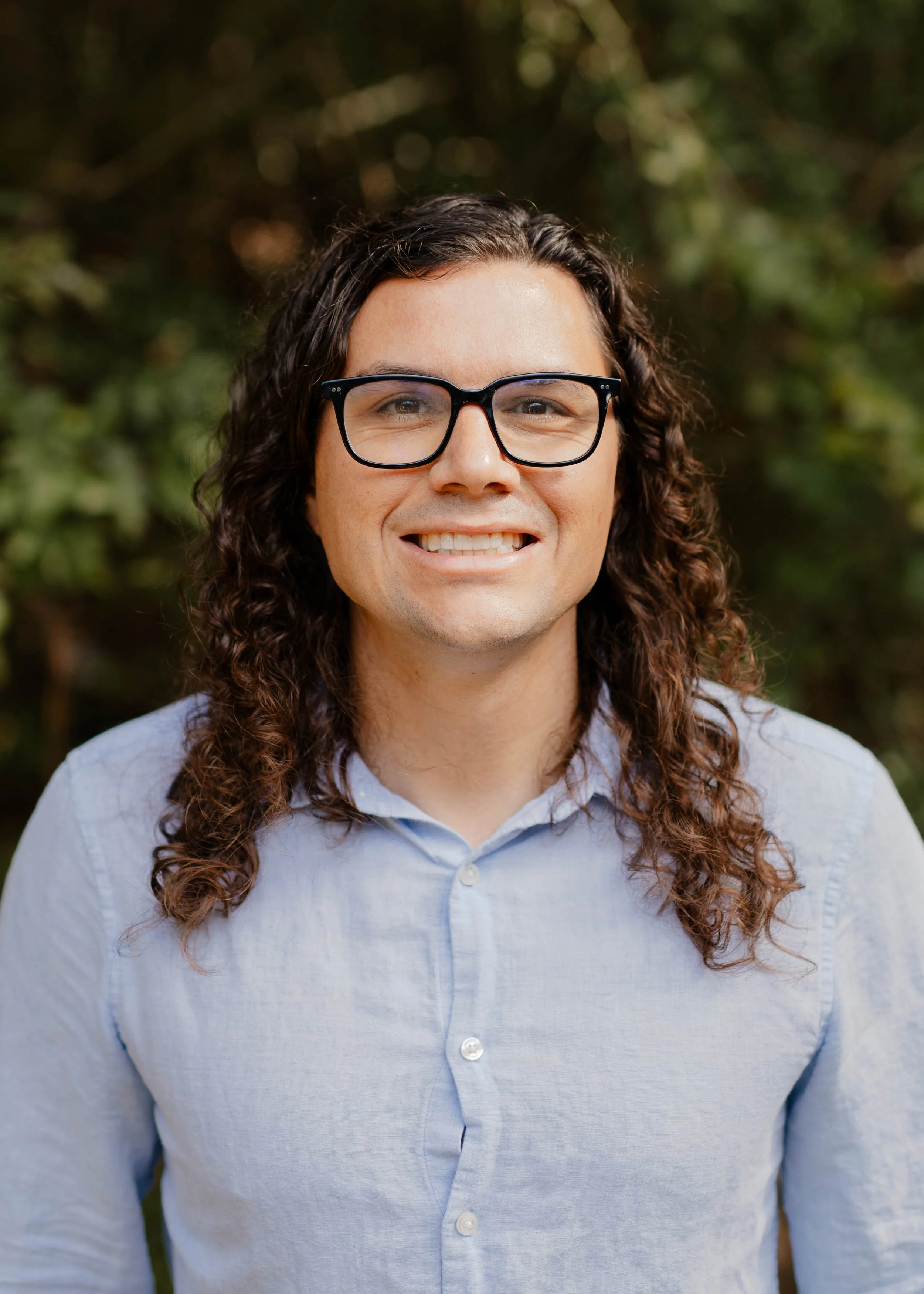 A man with long, curly brown hair and a serious expression, wearing a light blue shirt, outdoors during daytime.