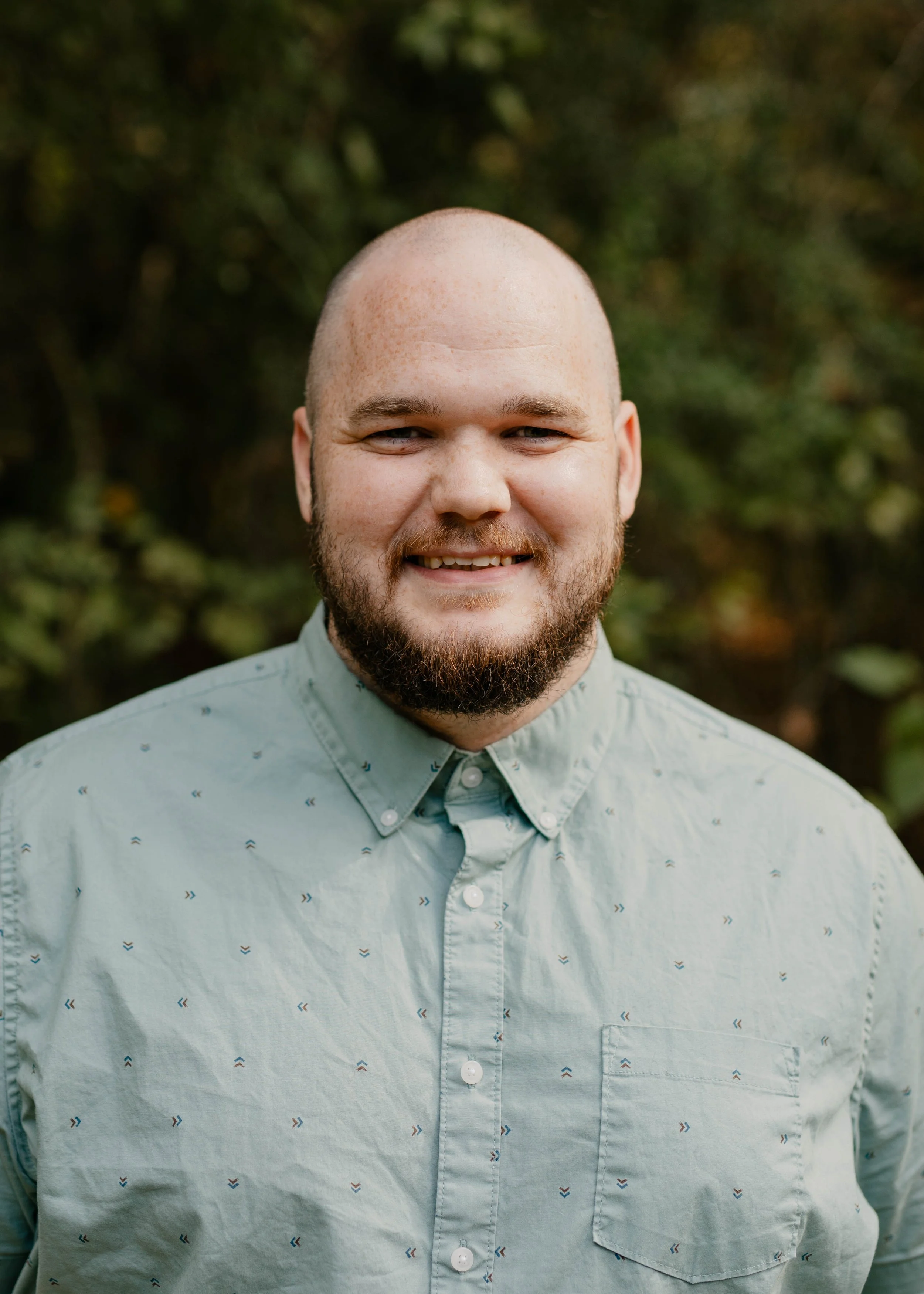 A man with a beard smiling, wearing a light blue collared shirt, standing in front of a beige wall with vertical panels.