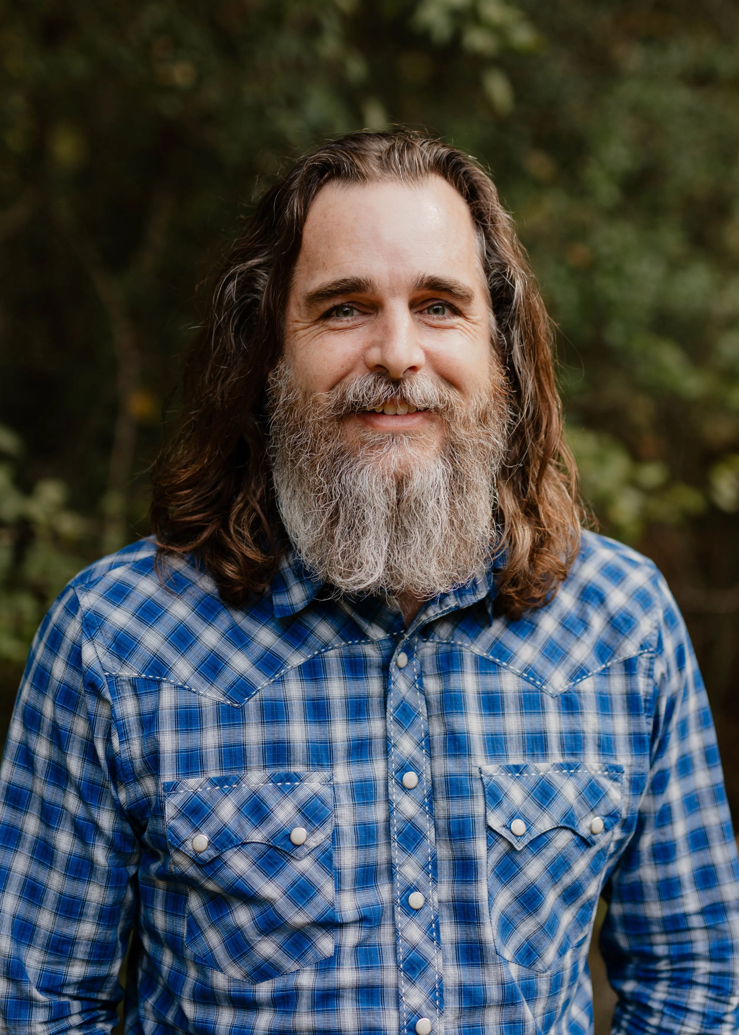 A man with a beard and mustache wearing a gray cap and a black collared shirt, smiling outdoors with trees and a multi-story residential building in the background.