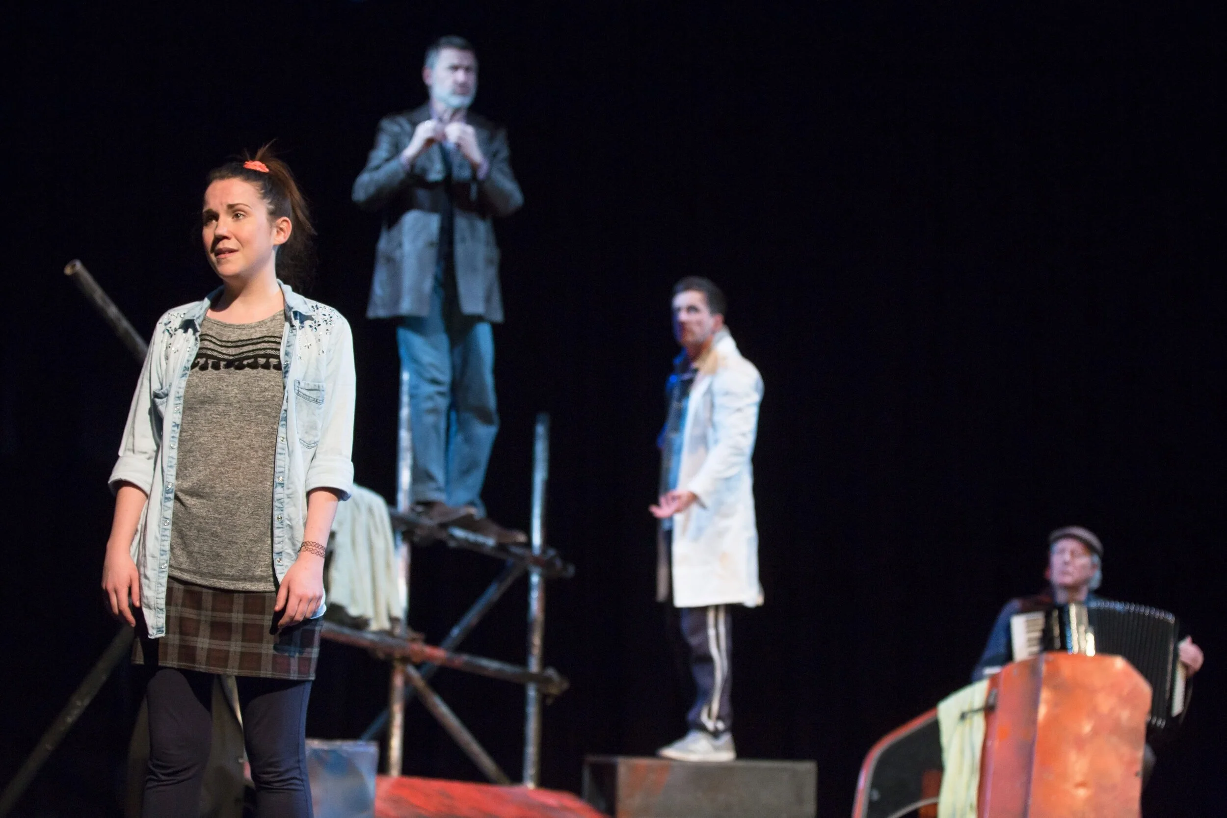 Four actors on stage during a performance of Into The West, with one woman in the foreground expressing emotion, and three men in the background, one on a scaffold, one standing, and one playing an accordion.