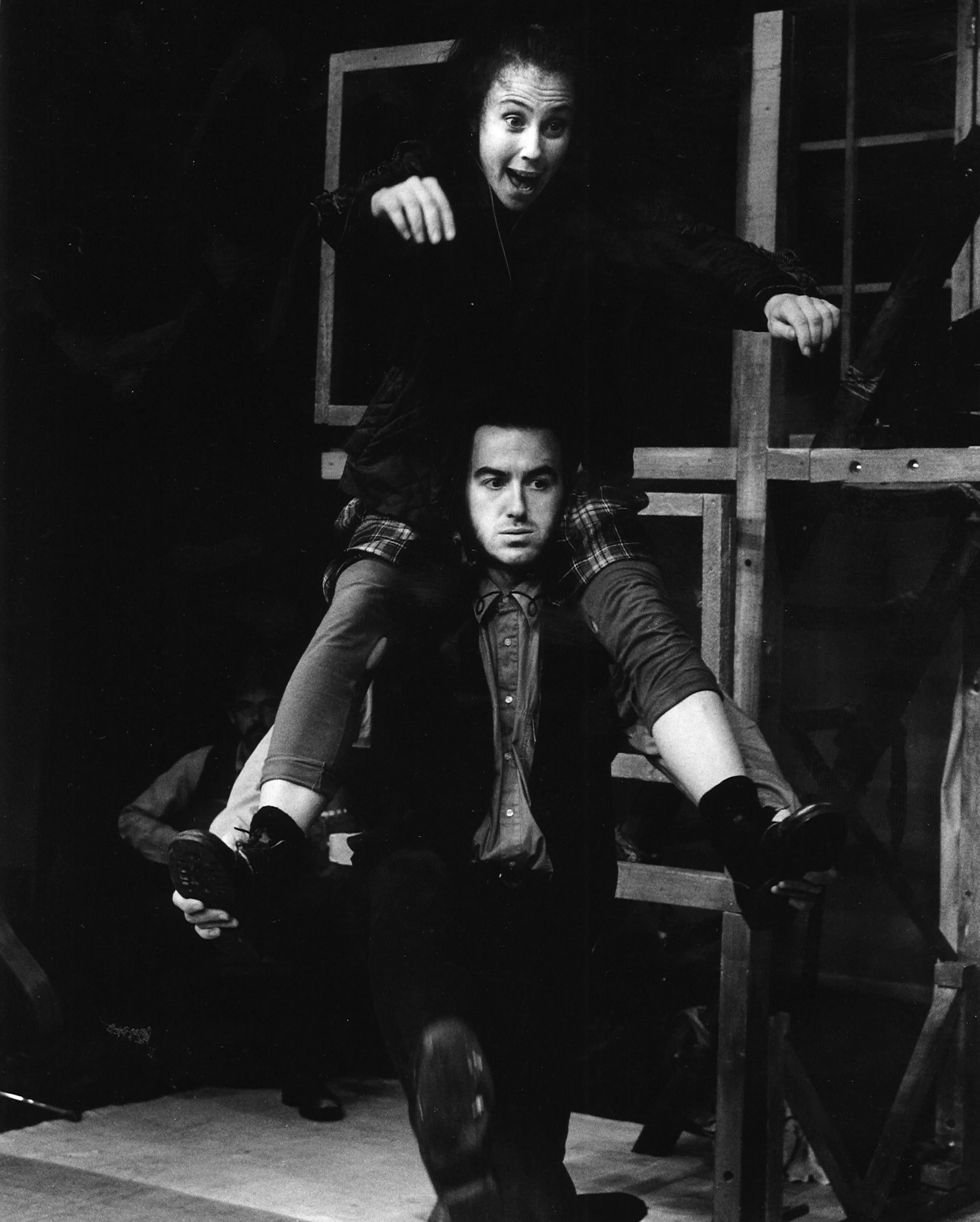 Black and white photo showing a woman with an excited expression sitting on a man's shoulders, who is walking forward with a serious look.