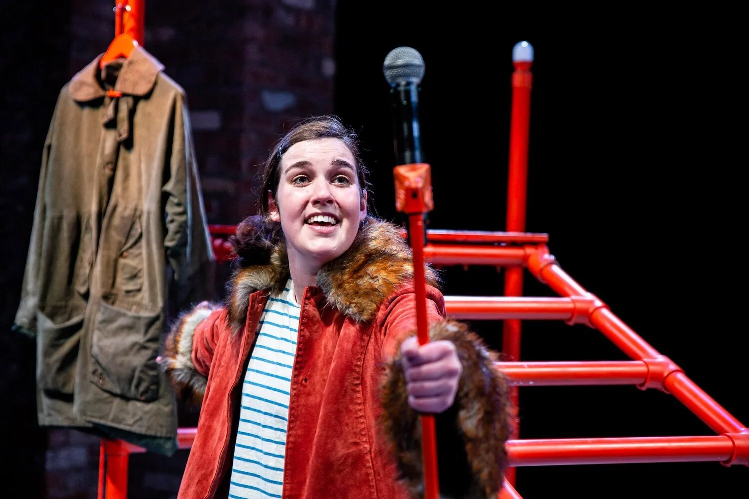 Safi looks forward towards the camera holding a microphone and stand in front of her.