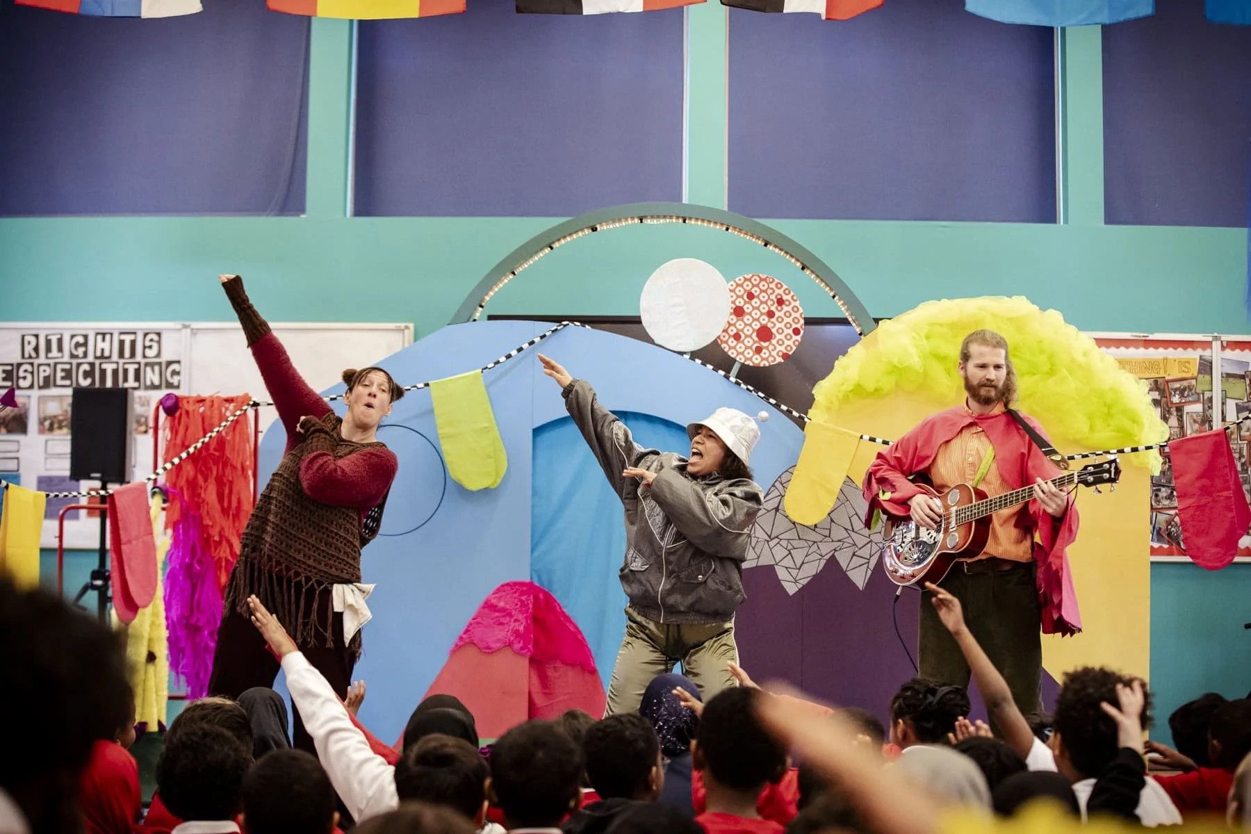 Three actors performing on stage. Two have one arm raised and one holds a guitar. Audience members in the foreground hold their arms raised too.