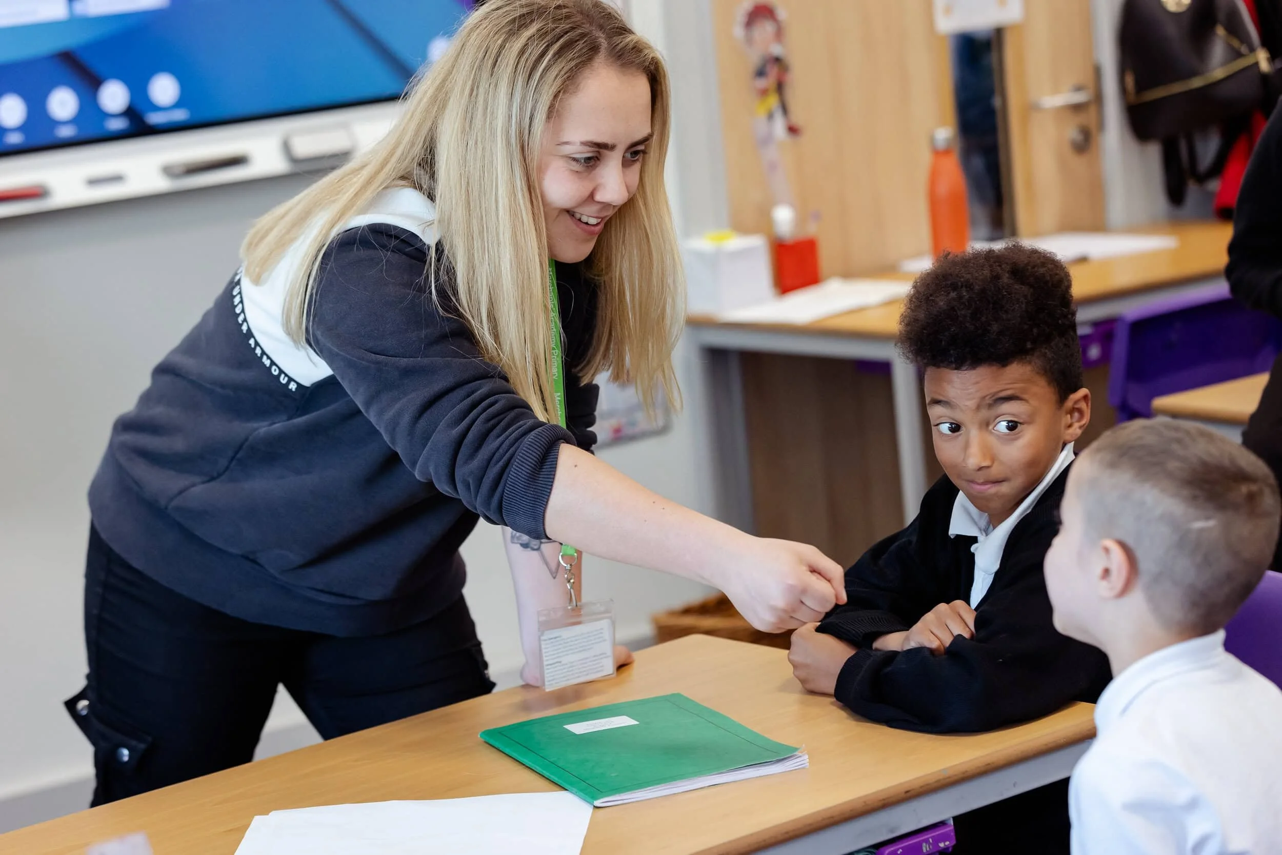 A teacher stands holding out a pretend microphone to a pupil who is sitting behind a desk and looking up at her. Another pupil sits next to them and looks on.