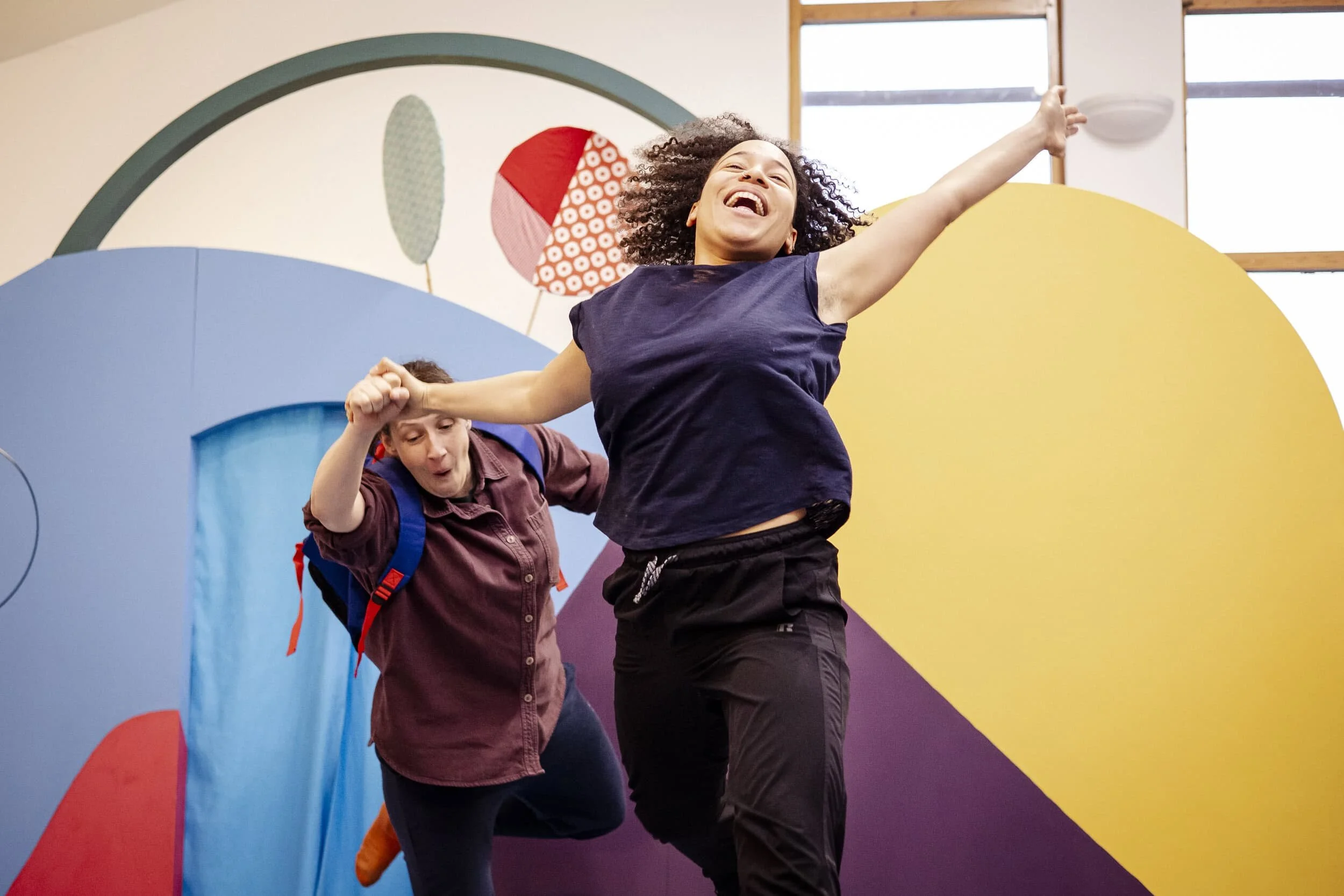 Remi and Amy performing in The Forest Door. Remi is in the foreground jumping with arms outstretched, Amy is behind hoding her hand and wearing a backpack.