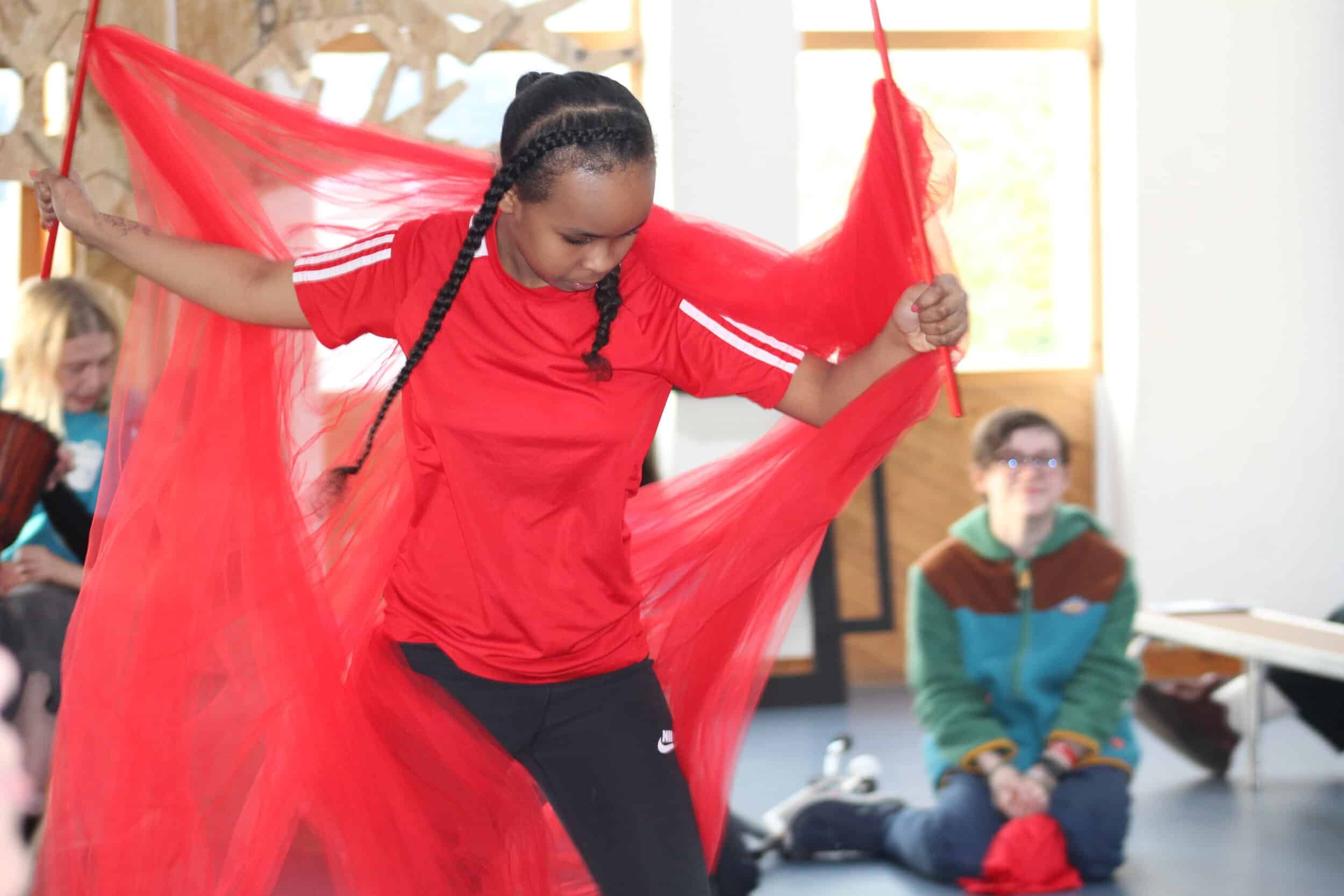 A member of Louder Than Words stands with arms outstretched holding two sticks with red material attached, which is flowing behind and around them.. They are wearing a red t-shirt with white stripes on the arms and black trousers..