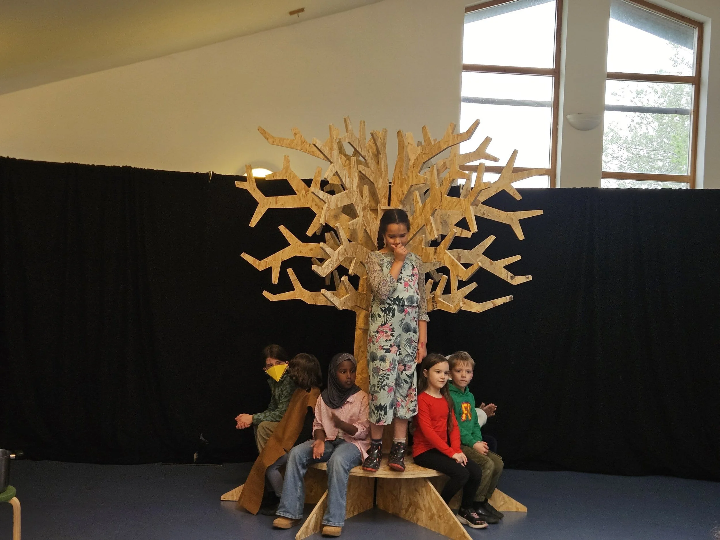 A member of 6-9's stands on a platform in front of a wooden tree, while 5 other members sit around the tree. Behind them is a black curtain.