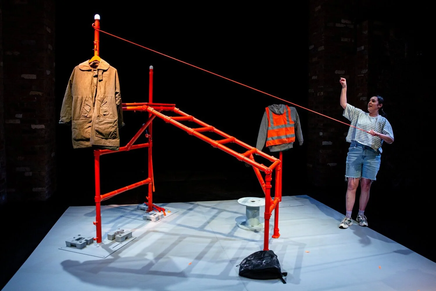 Saffi stands stage right pulling a cord that pulls up the rain man's coat. On the stage is an orange metal frame.