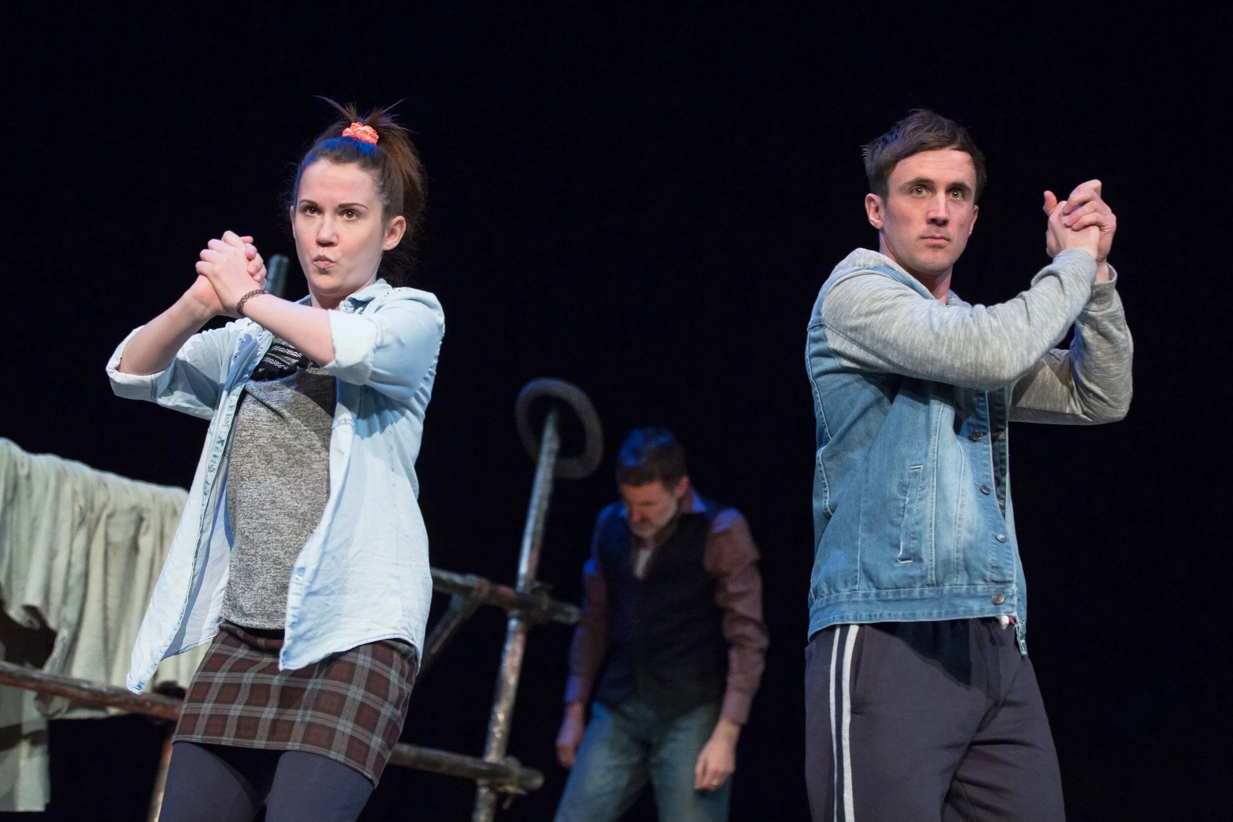 Three actors on stage during Into The West. The woman on the left is wearing a light denim shirt over a grey top, plaid skirt, and dark tights, with her hair up in a ponytail secured with a pink scrunchie.