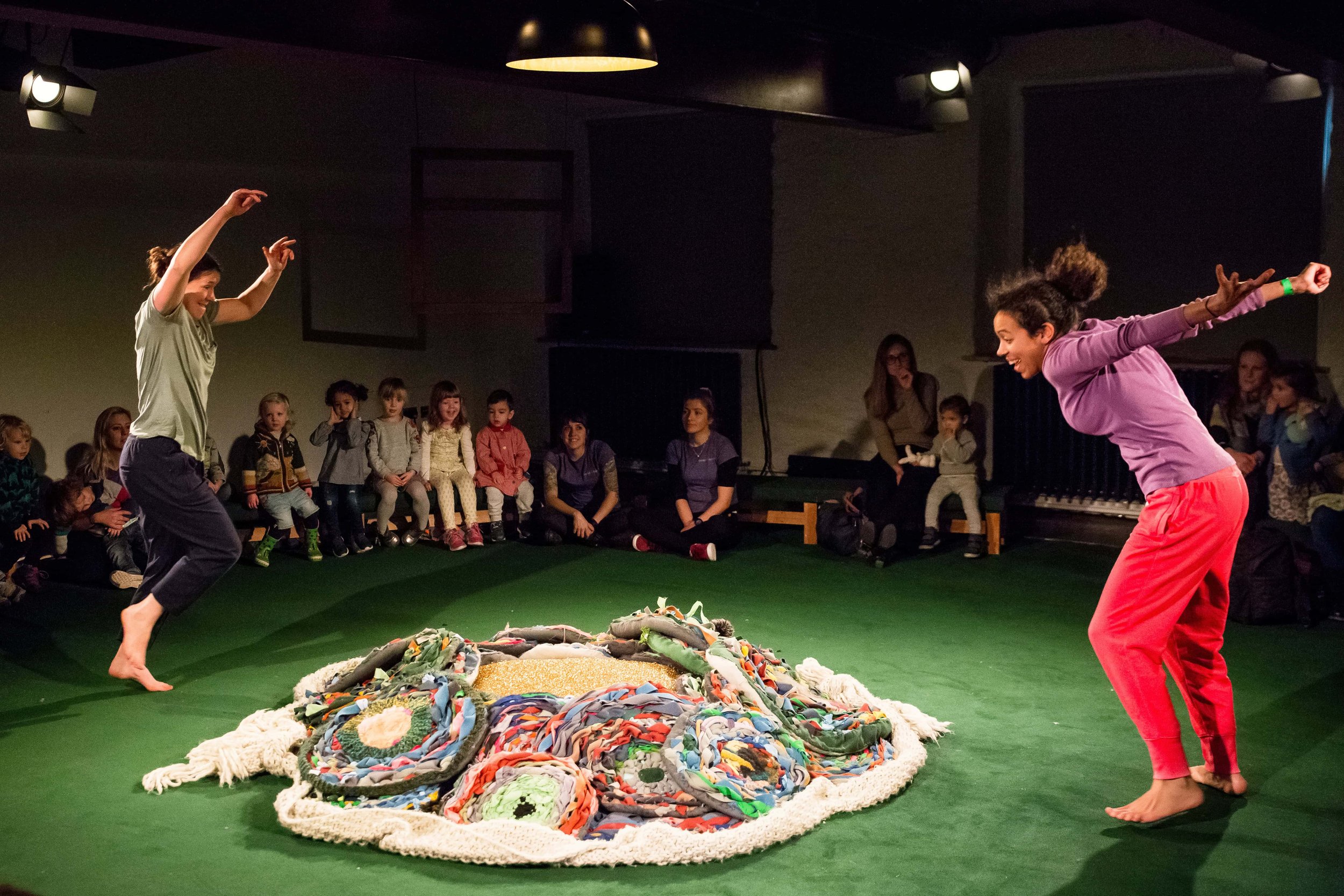 Two performers stand either side of a pile of colourful cicle mats and are about to jump onto them. Audience members look on in the background.