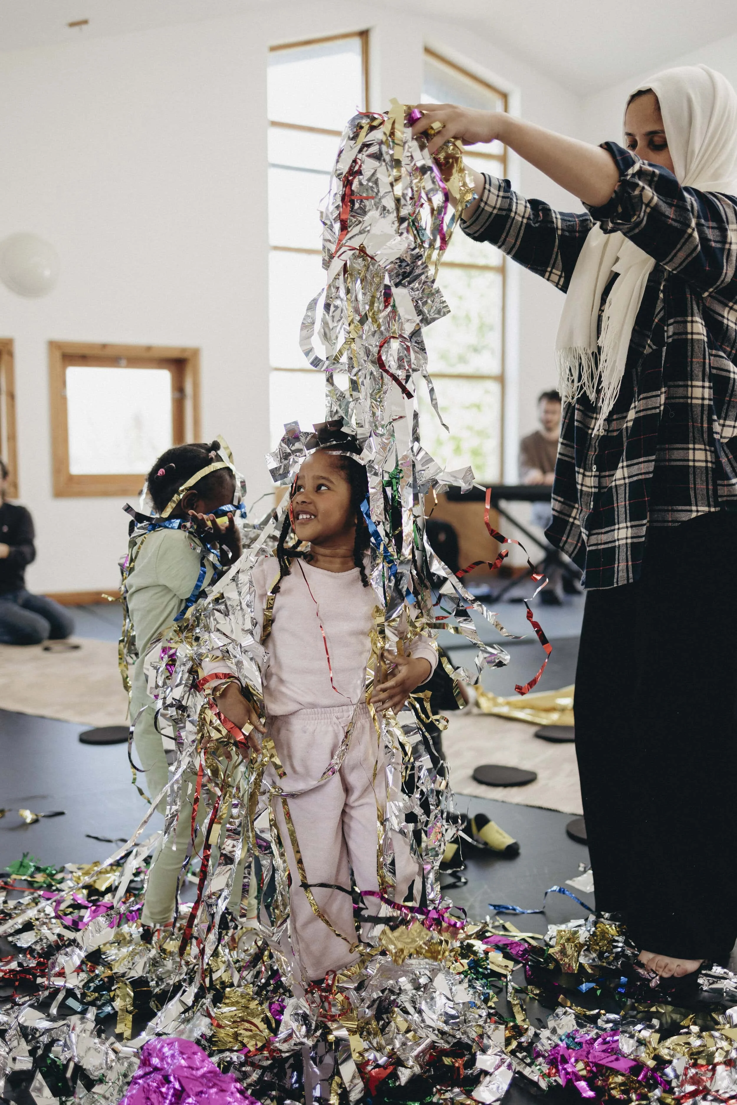 Two young children stand with colourful ribbon on and around them, which is being dropped by an adult from above them. 