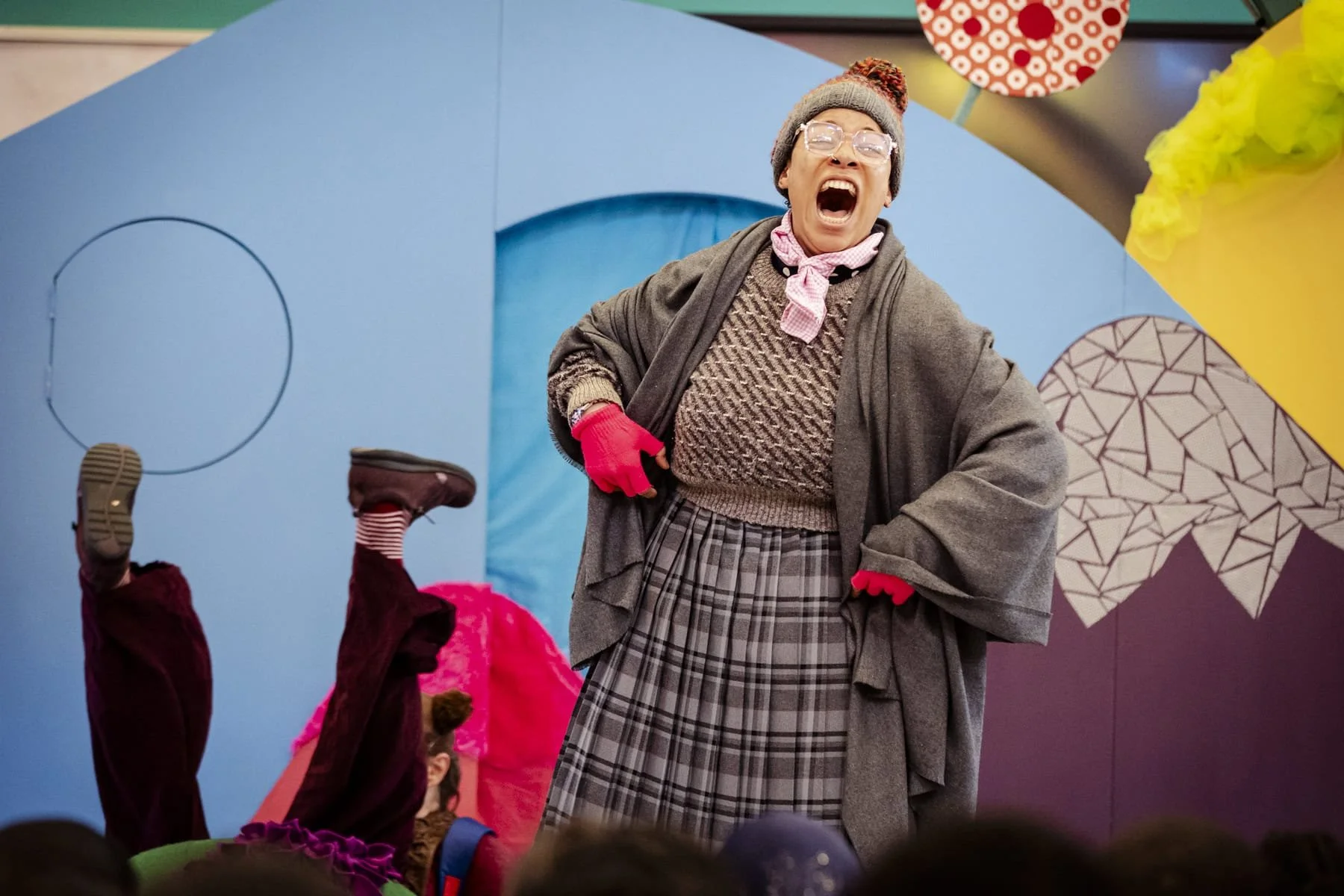 A woman dressed as an old lady with glasses, a knitted sweater, a plaid skirt, and a grey coat, shouting loudly. She is wearing a grey beanie with a pom-pom and pink gloves.