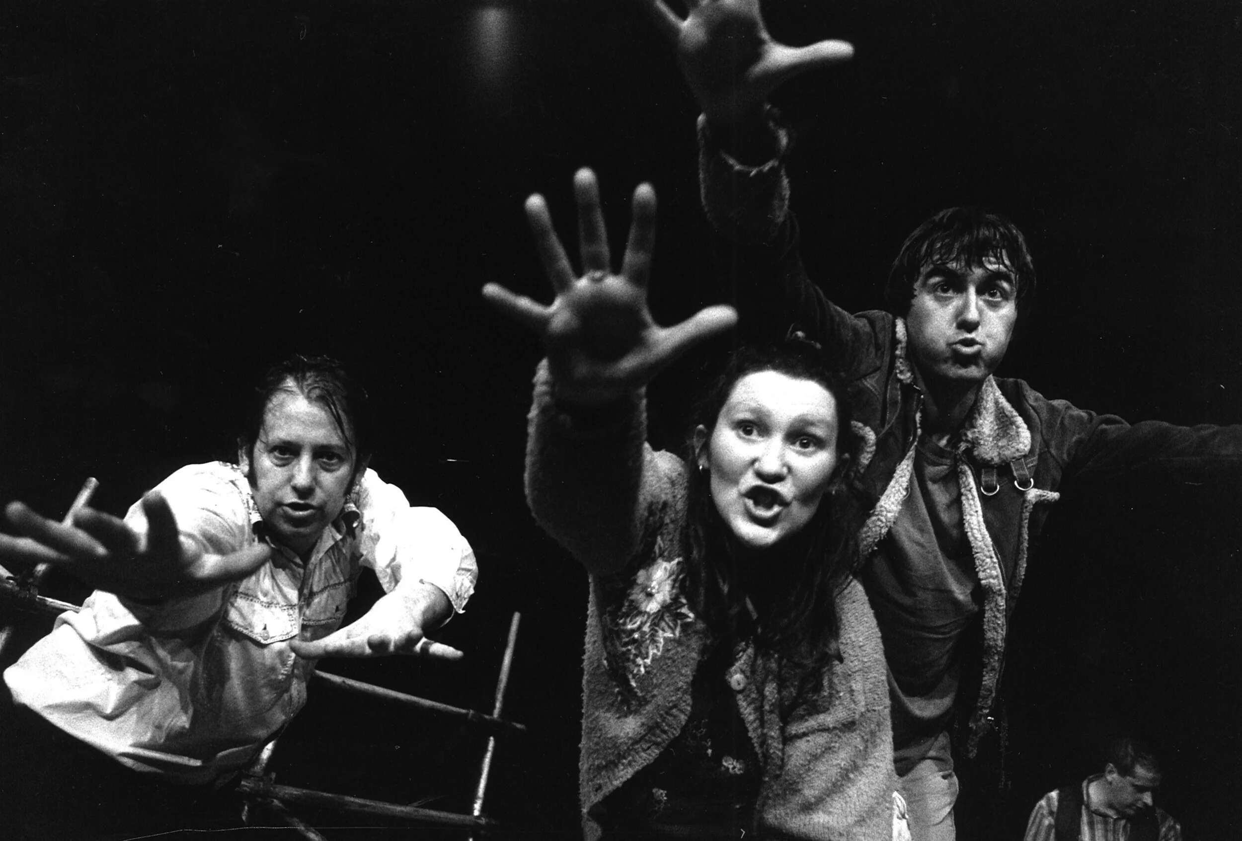 Black and white photo of two adult performers pointing and reaching towards the camera with an intense expression, with a third person in the background.