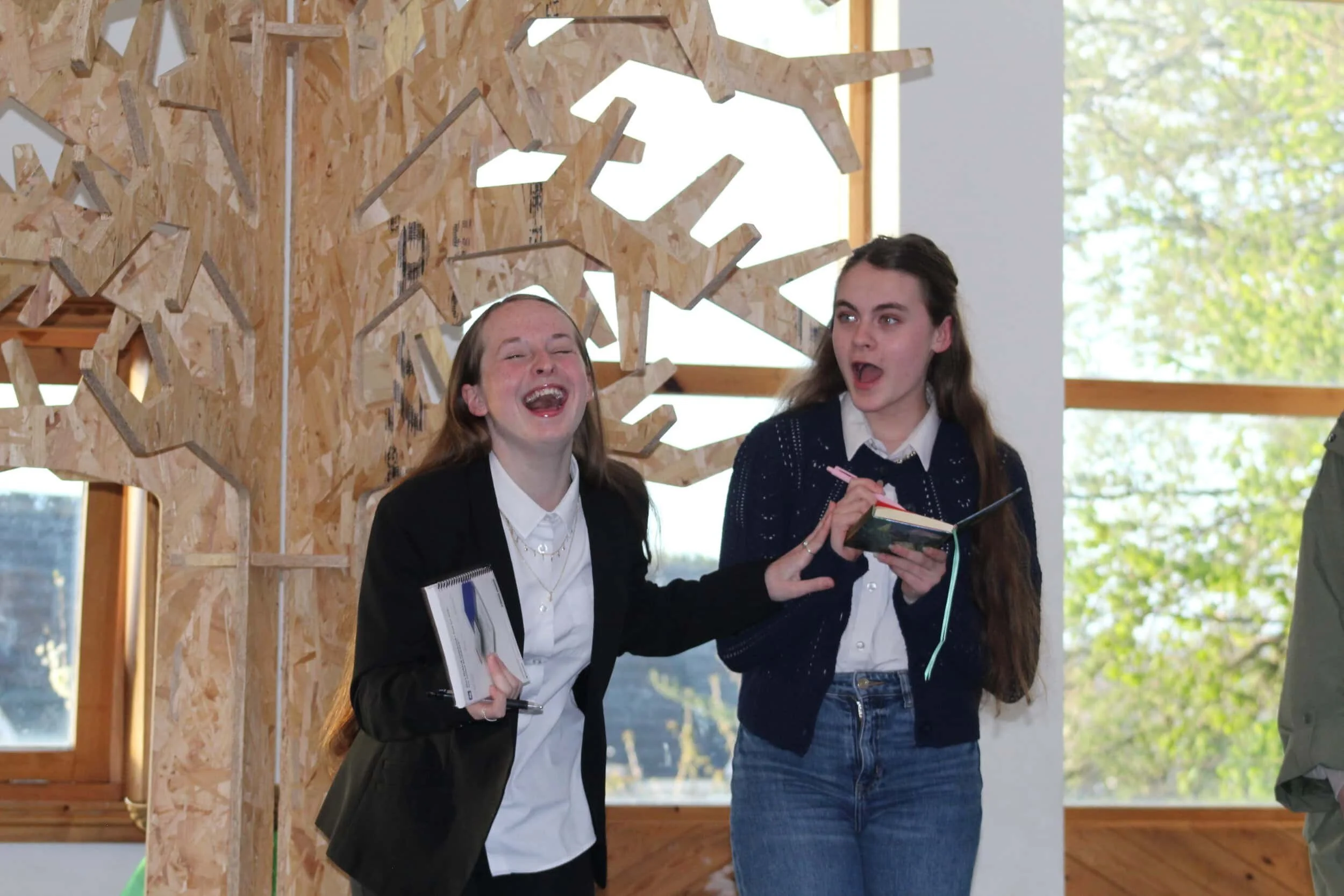 Two members of Louder Than Words stand laughing in front of a large wooden tree. One is holding a pen and writing into an open book.