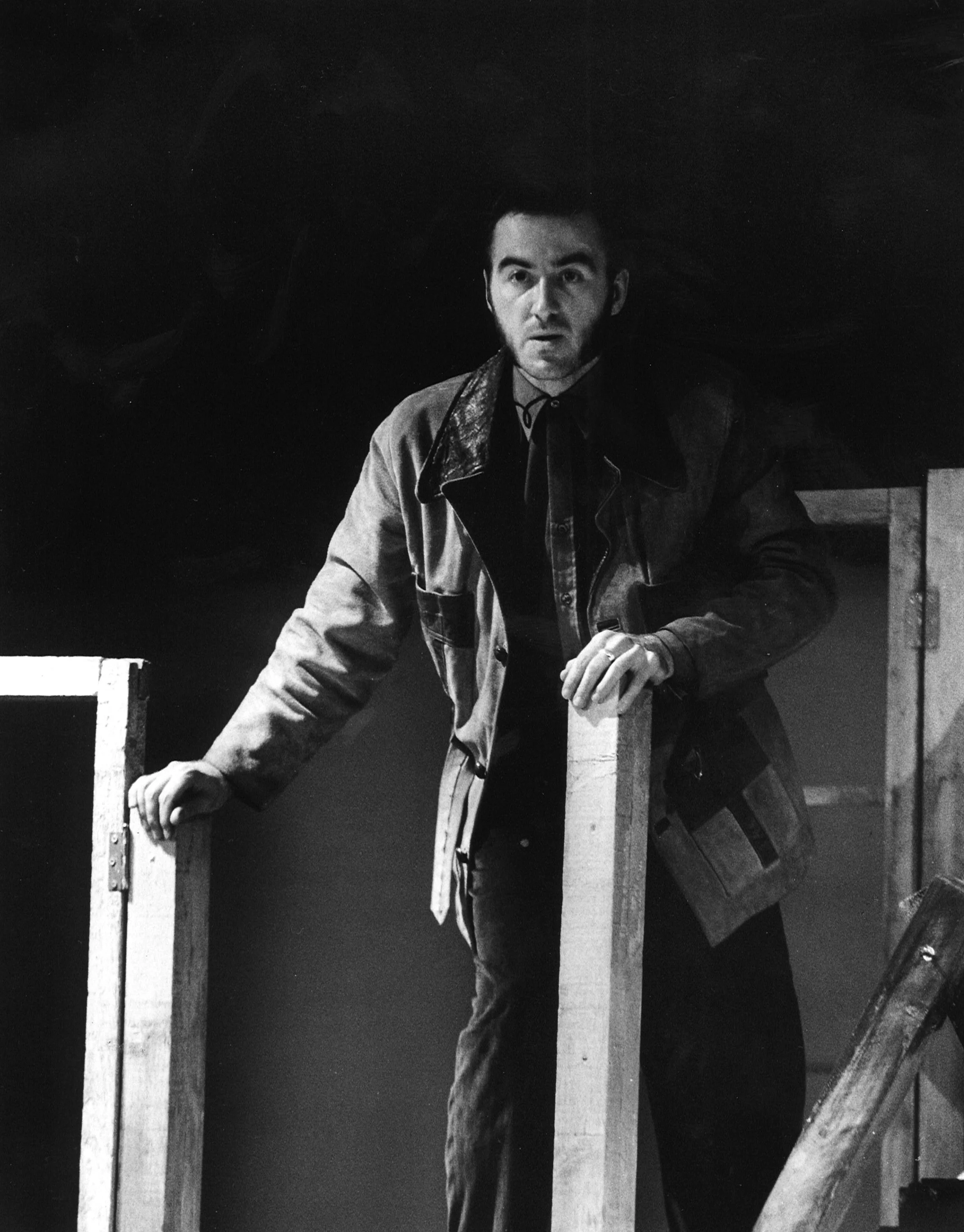 Black and white photo of a man with dark hair and beard, wearing a jacket, standing behind a wooden railing on a stage.