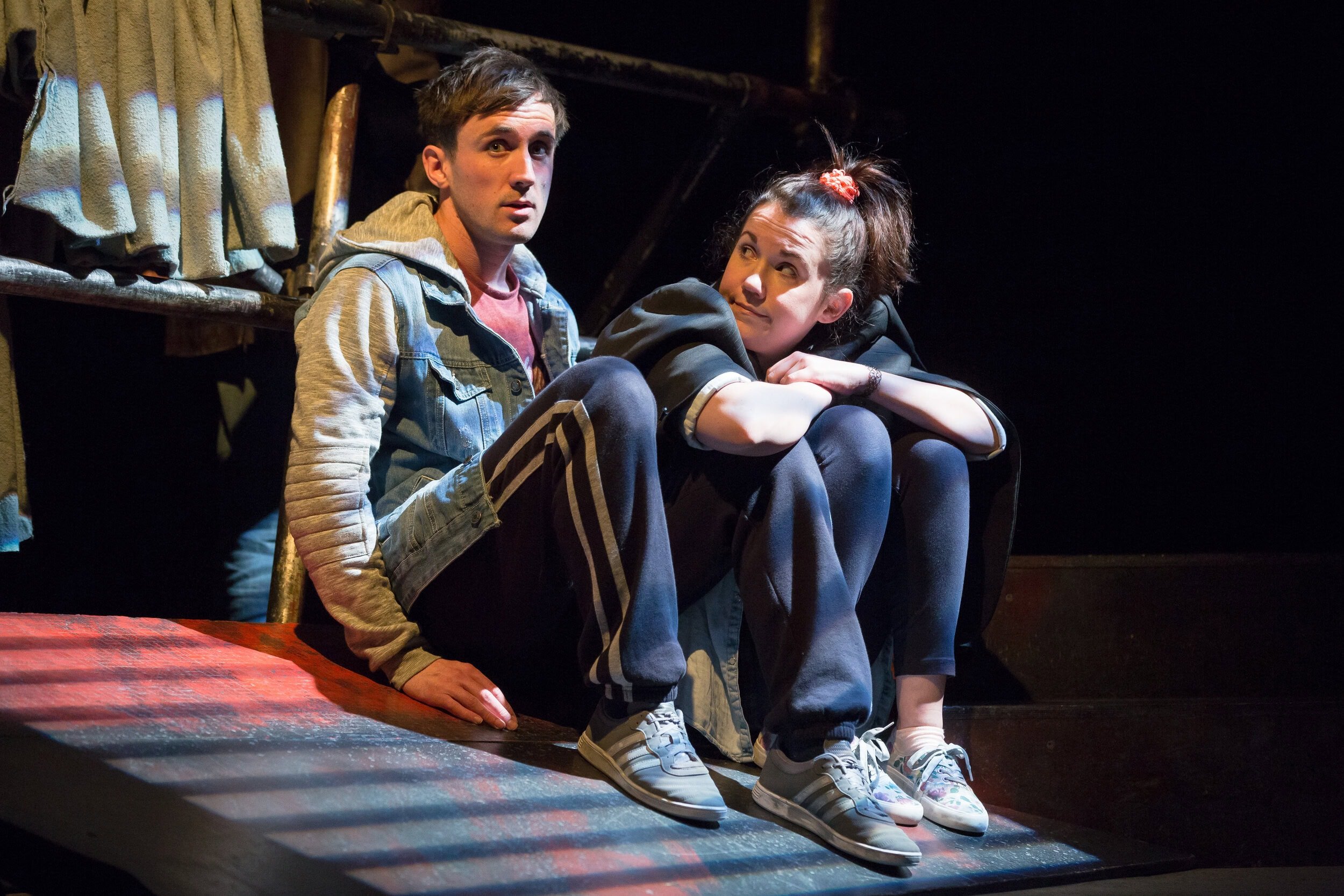 Two young people sitting on a stage, one with a denim jacket and another wearing a black hoodie, both looking to the side during performance of Into The West.