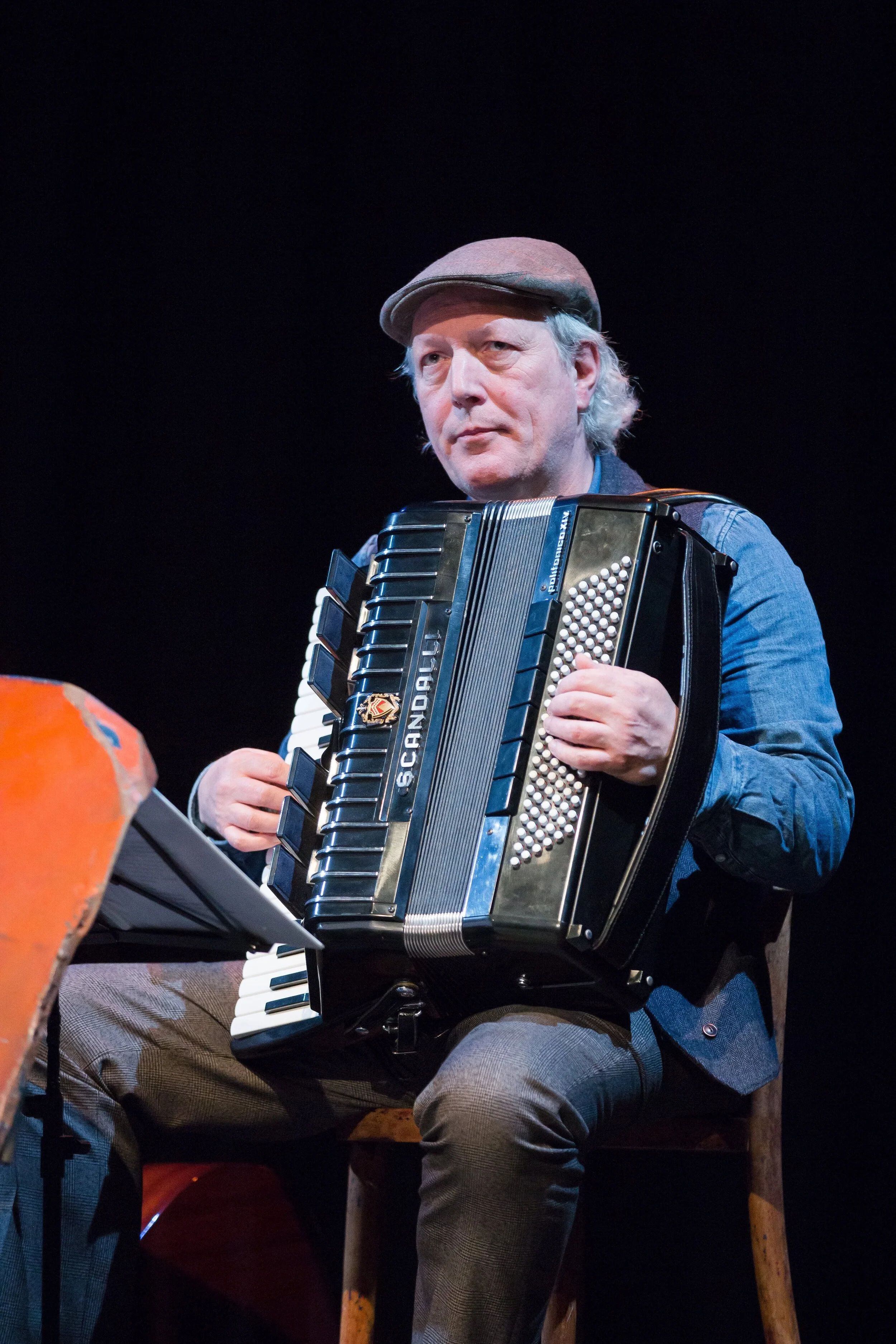 An older man wearing a grey cap and a blue shirt sitting on a wooden chair on stage, playing an accordion.