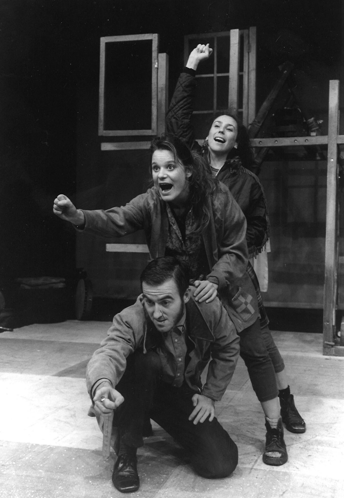 Three actors on stage, performing in Into the West. One actor is kneeling and pointing forward, another is standing on their back, and the third is raising a fist in the air. The background features stage set pieces.