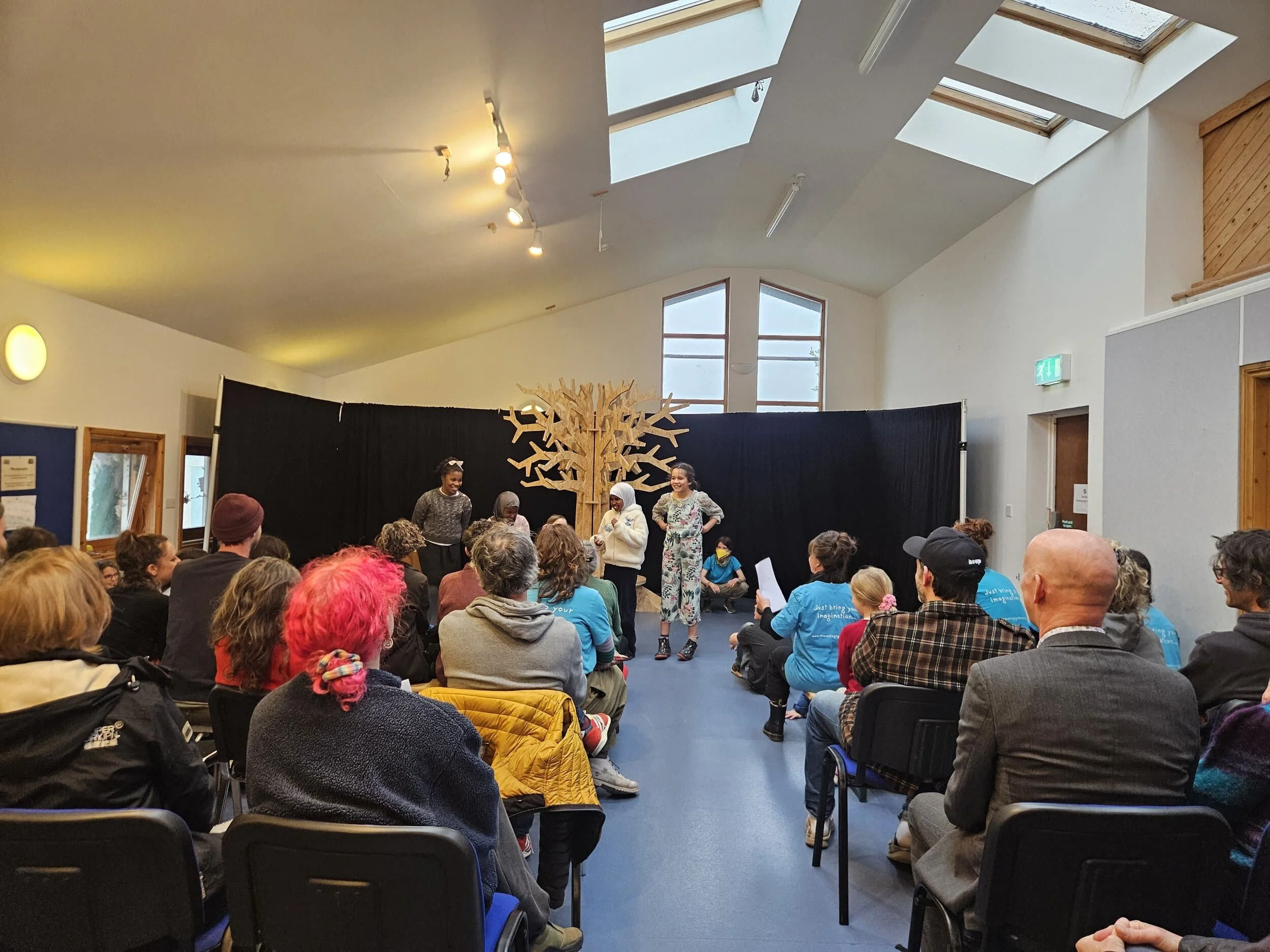 Members of 6-9's perform in front on an audience. Four children stand in a line in front of a wooden tree, with audience members sitting in rows in the foreground.