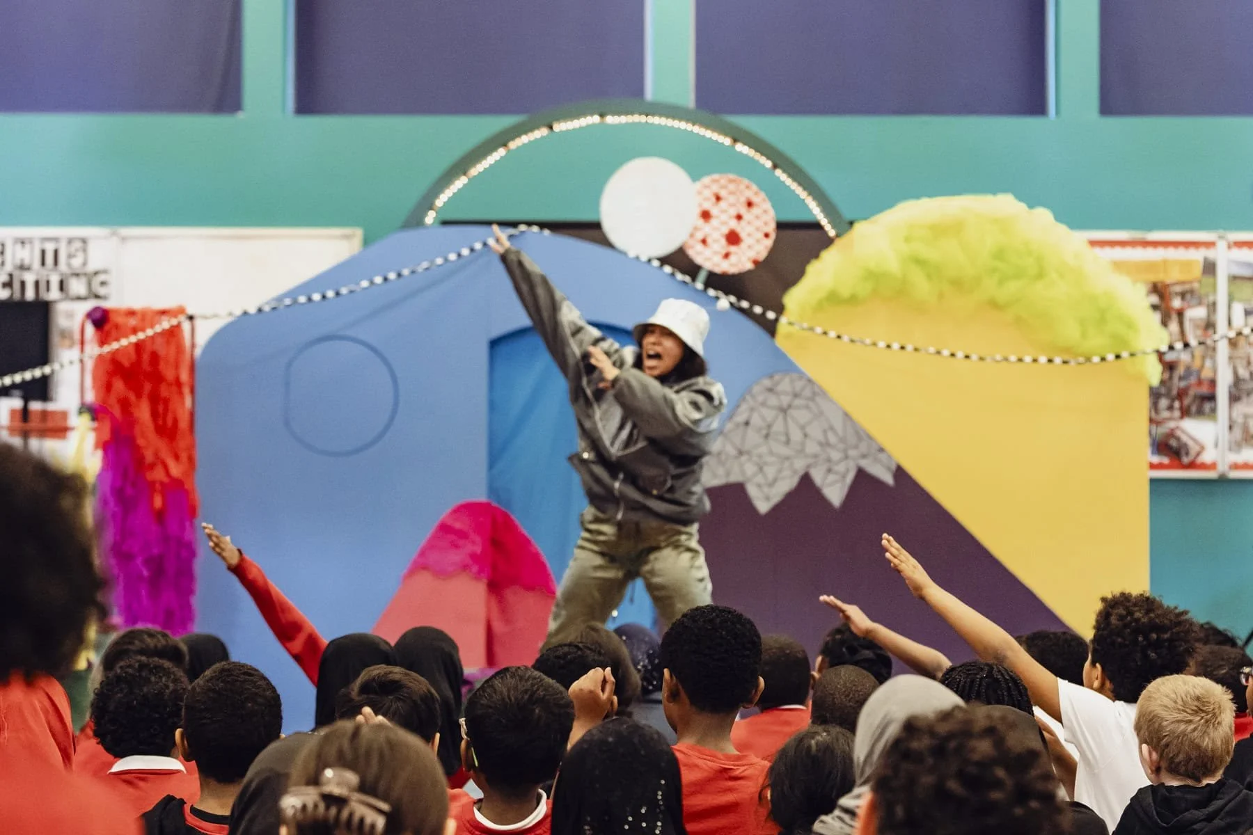 An actor on stage wearing a white bucket hat and camouflage pants raises their arms. Pupils sit in the foreground watching the performer and imitating her movements.