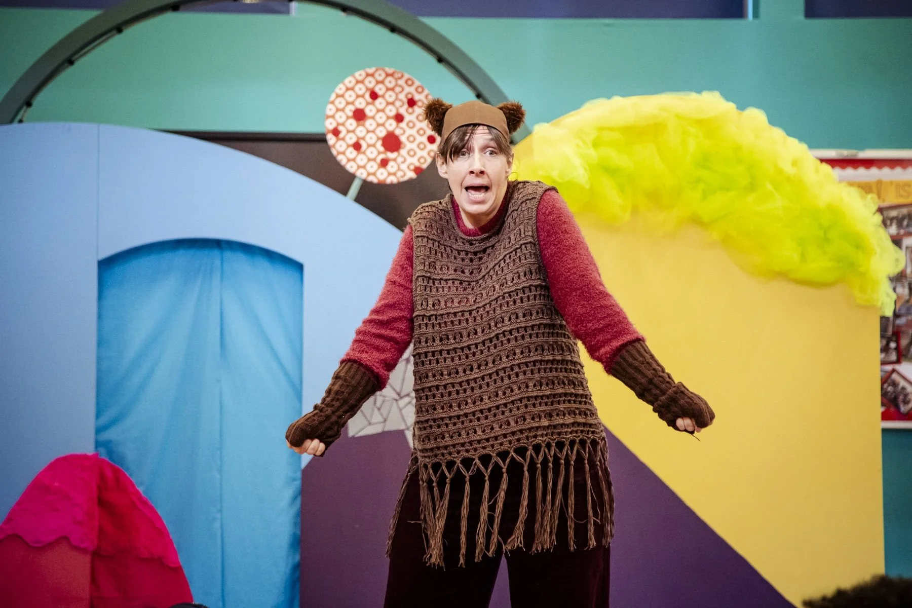 A woman with a surprised expression is wearing a bear-ear hat and a crochet top. She is standing in front of a colourful set with large shapes.