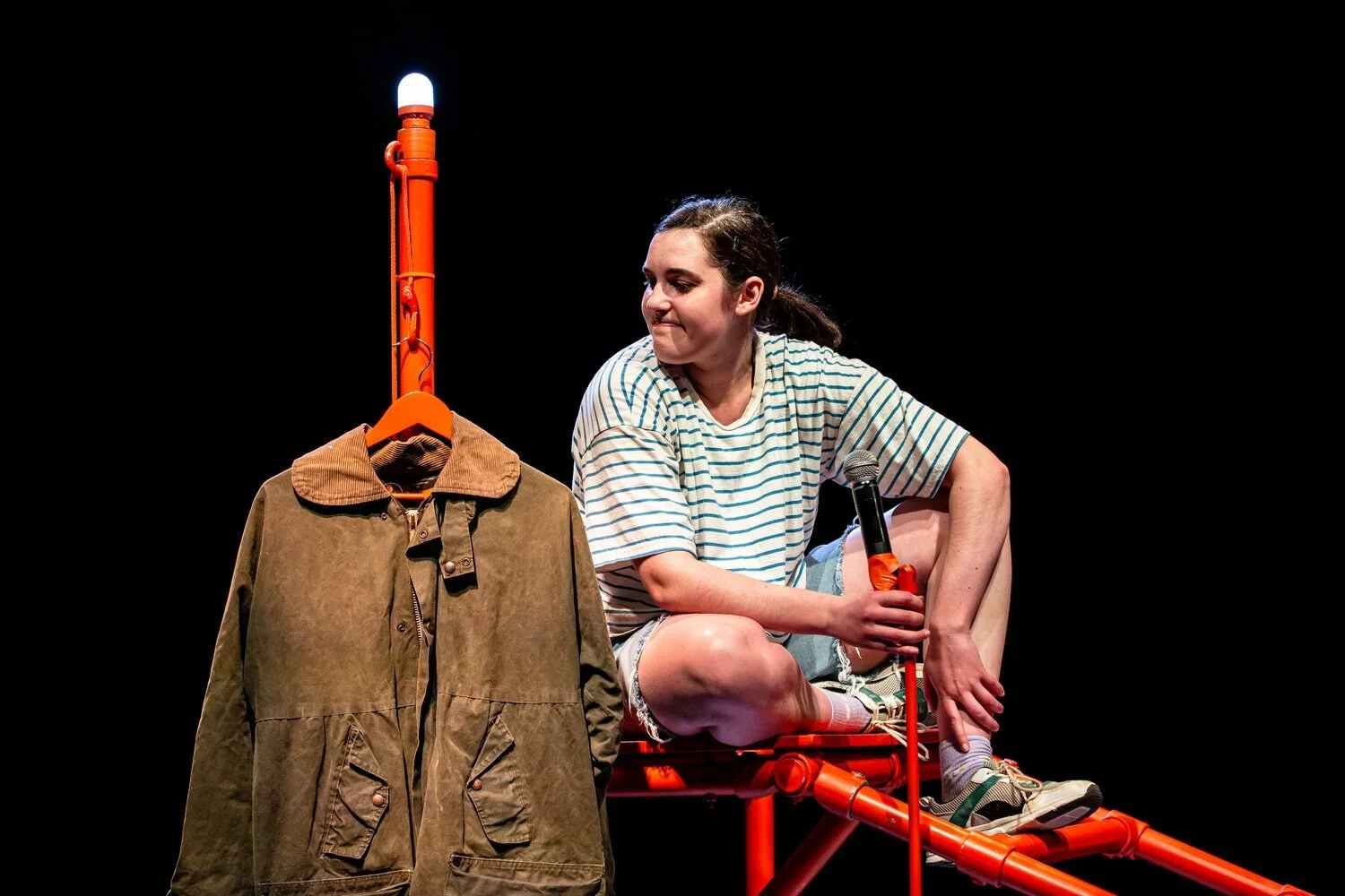 Saffi sits on the orange frame with microphone in hand and looking towards the rain man's coat.