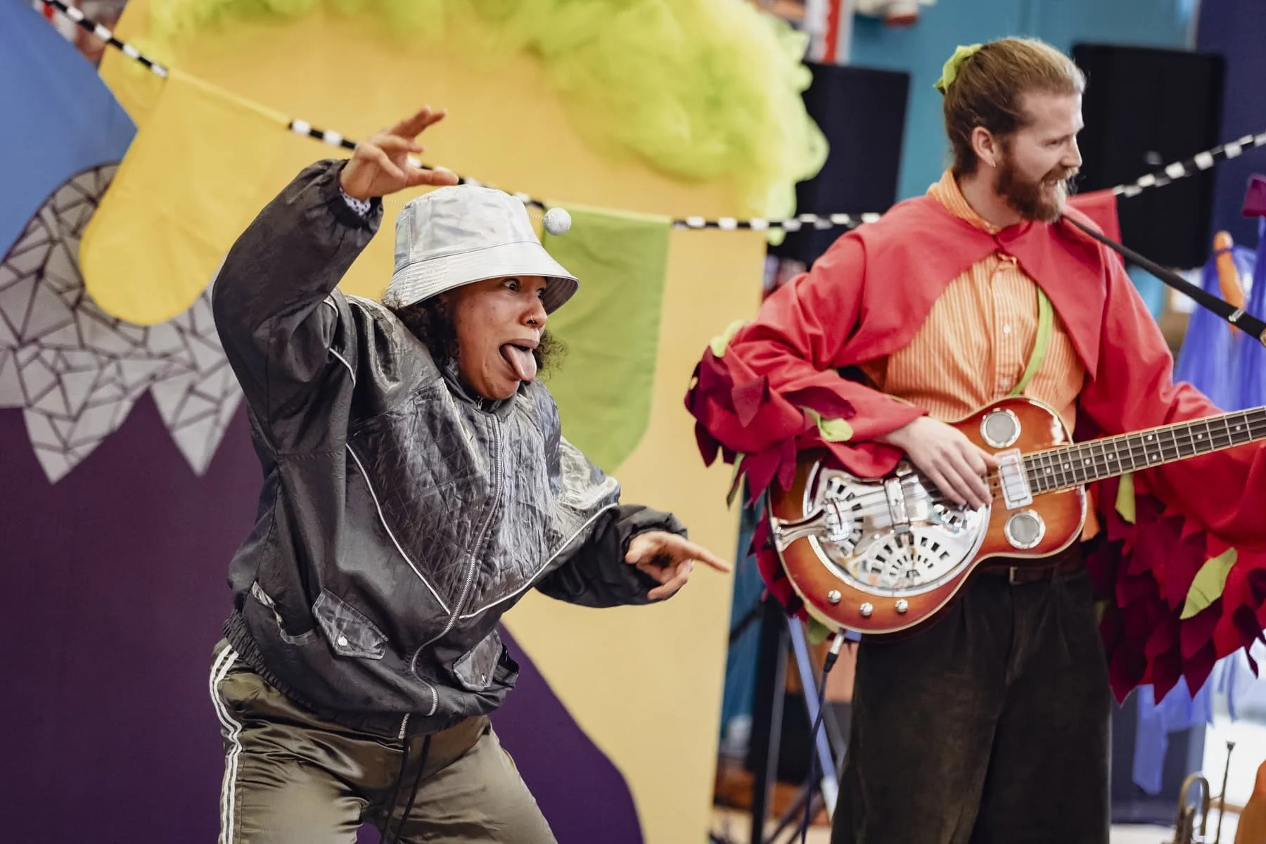 Two actors performing on stage, one person wearing a grey bucket hat and jacket making a funny face with tongue out, and the other person playing a guitar, wearing a red cape, smiling.