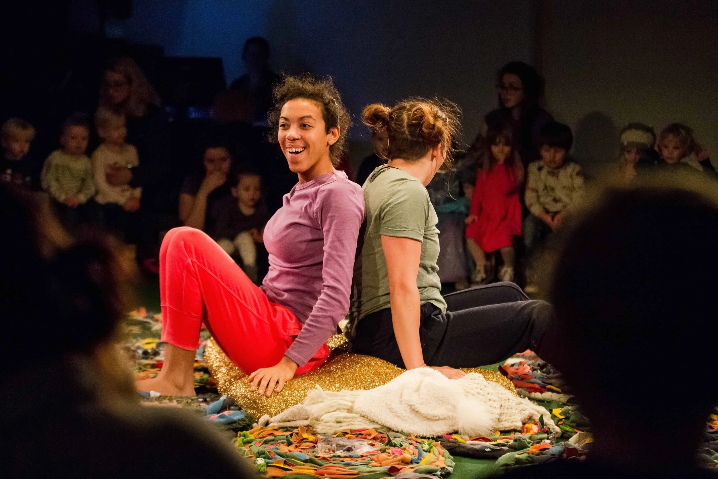Two perfomers sit on a gold cushion with backs together facing in opposite ways.