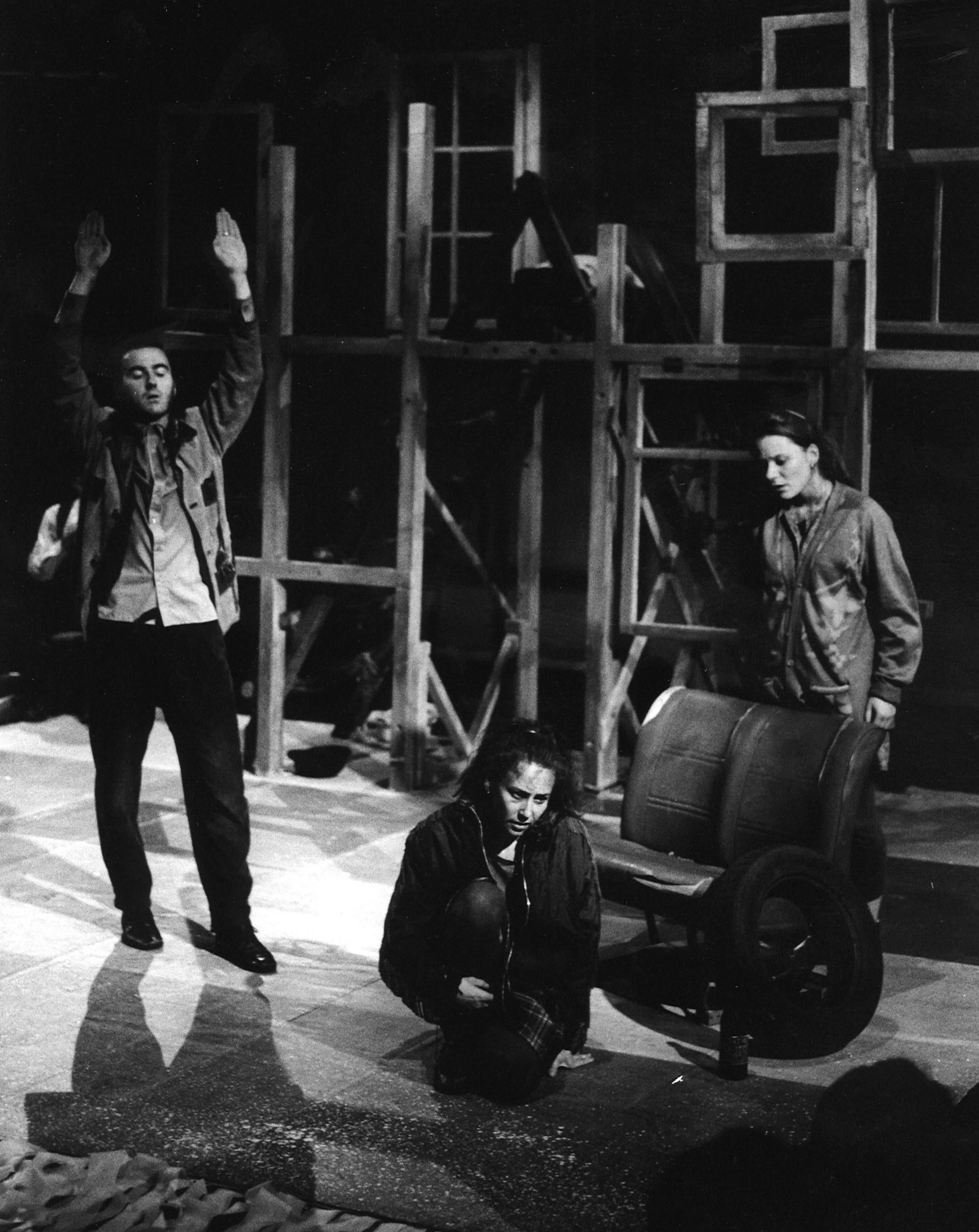 Black and white photo of three actors on stage, one sitting on the floor holding their knee, another standing with a suitcase, and the third raising their hands, with a wooden set behind them.
