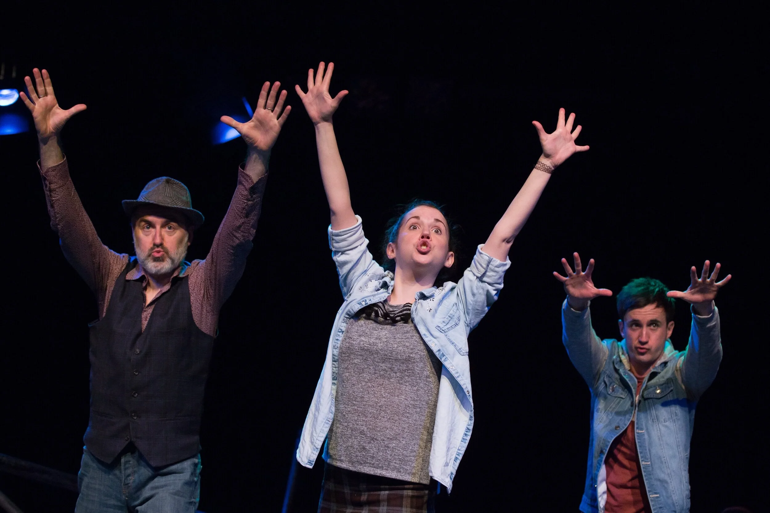 Three actors on stage performing with their arms raised and making expressive facial gestures, set against a dark background.