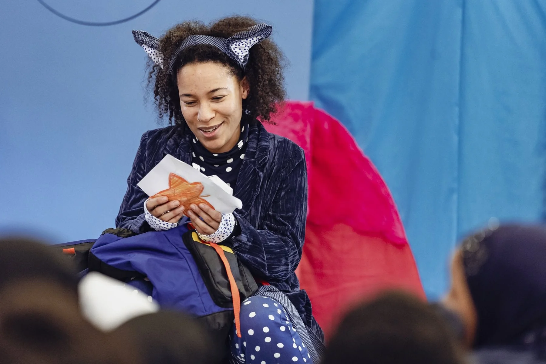 Young woman with curly hair, wearing a polka-dot bandana, polka-dot shirt, and a navy jacket, sitting and reading leaves that children have written, with a blue backpack on her lap, in front of a blue set.