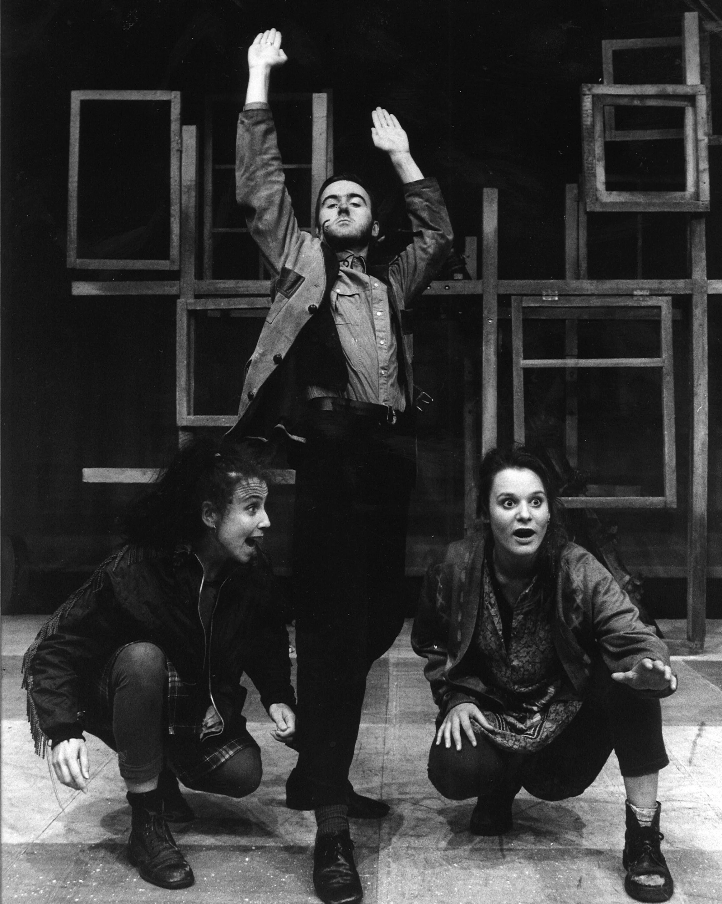 Three actors performing on stage, with two crouching and one standing. The standing man has his arms raised, and the two other actors are looking up at him with expressive faces.