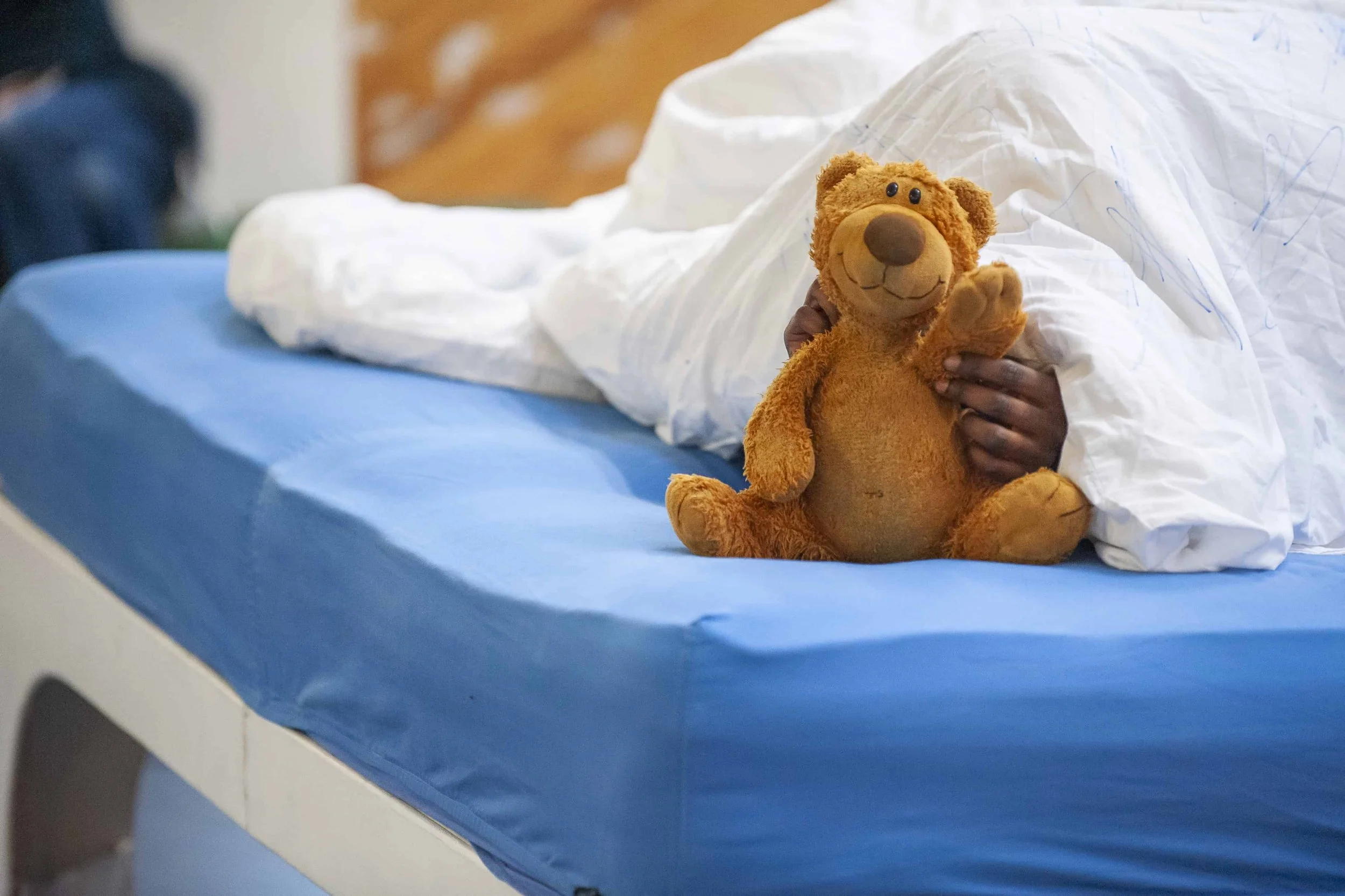 A small brown teddy sits on the blue mattress waving towards the camera.