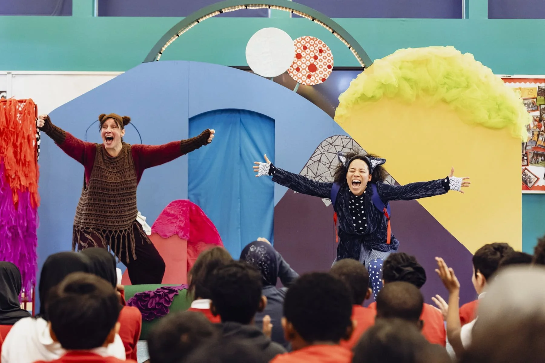 Two performers with expressive faces and outstretched arms perform to pupils in front of a colourful set. Audience members raise their hands in excitement.
