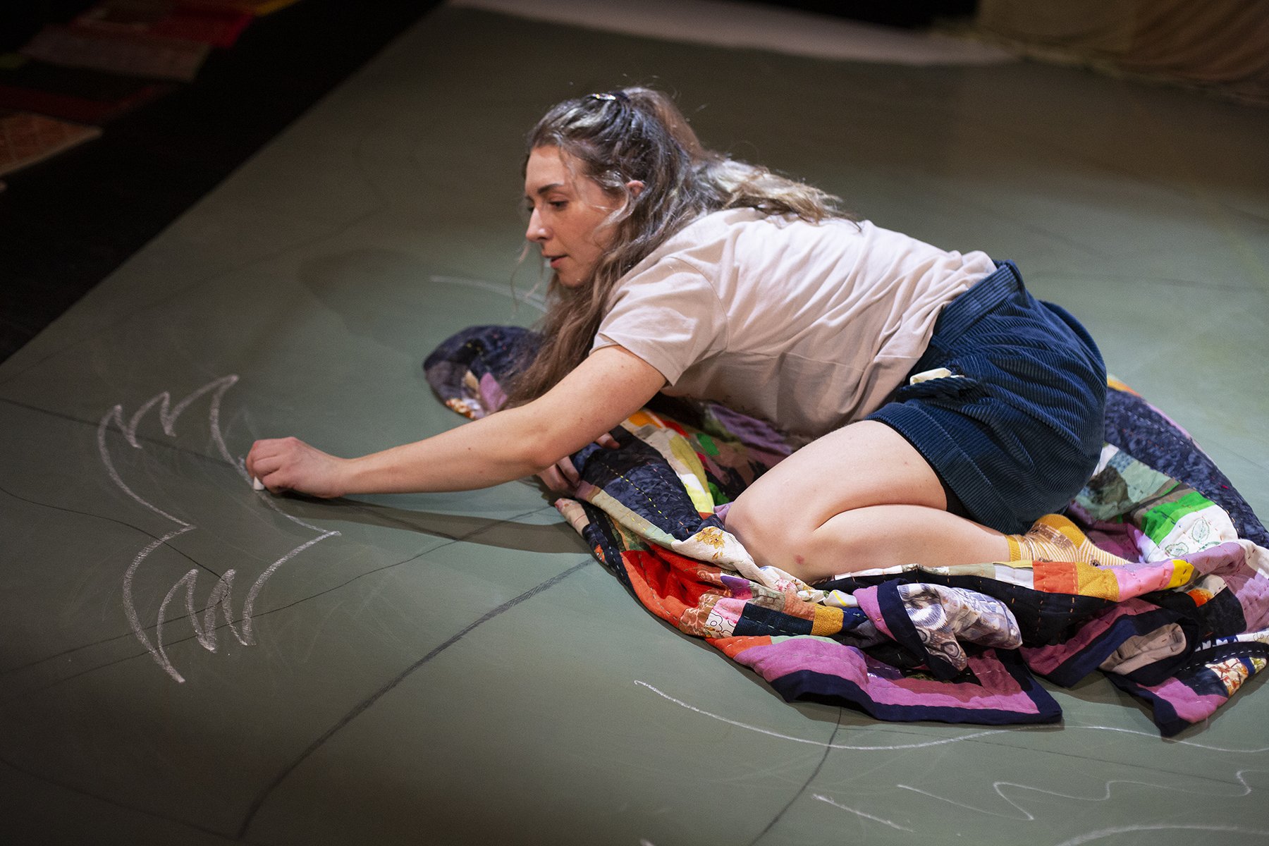 A woman with long brown hair in a ponytail, wearing a beige T-shirt and black shorts, is lying on her side on a green floor, drawing with chalk on the surface.