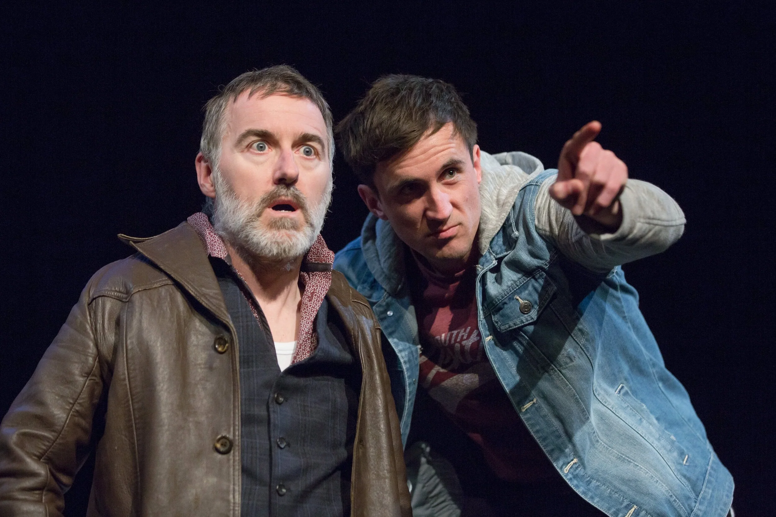 Two men are on stage, one older with grey hair and beard wearing a brown leather jacket, expressing surprise. The other younger man with dark hair wearing a denim jacket is pointing at something.