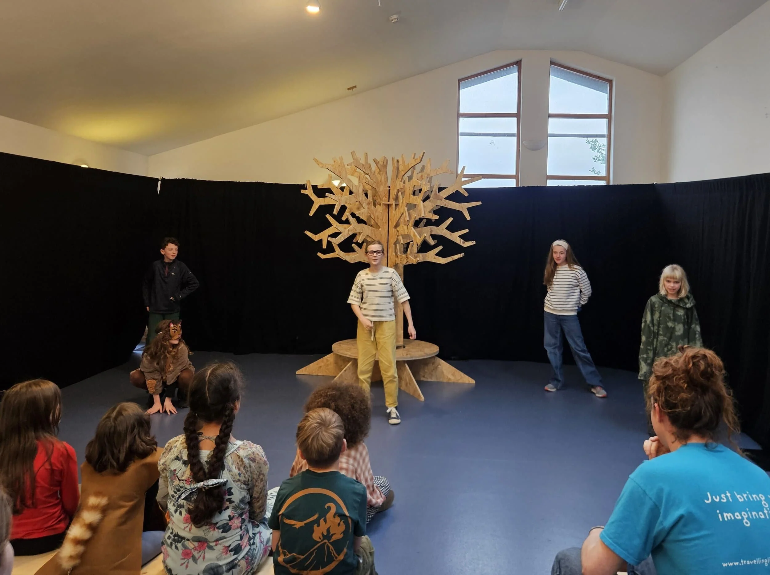 Four members of 10-12's Youth Theatre stand on stage with a wooden tree behind them and a black curtain behind the stage. A 5th member wears a cat mask and crouches on the ground. In the foreground a seated audience watches the actors on stage.