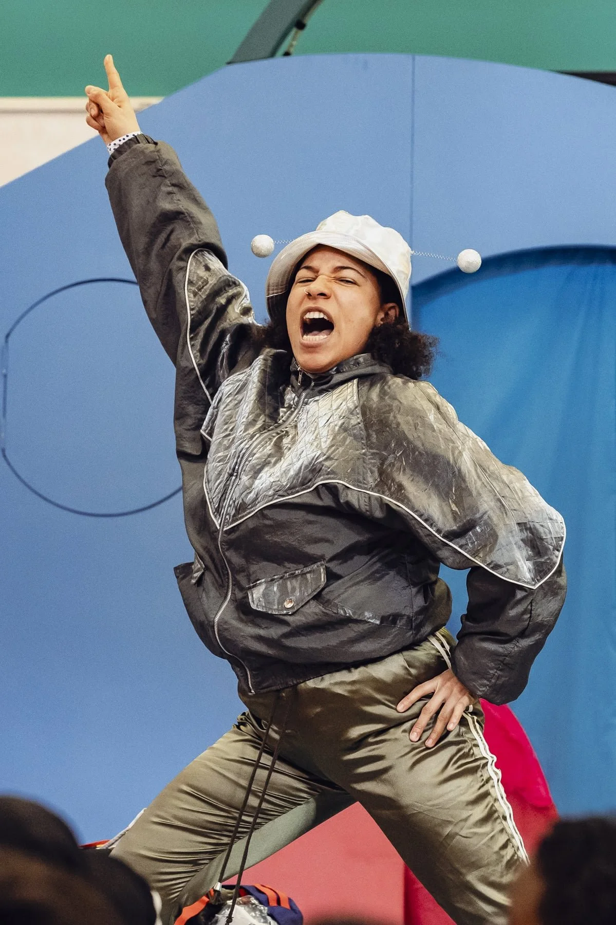 A woman in a dynamic, expressive pose with her mouth open and her right arm raised, pointing upward. She is wearing a light-colored bucket hat, a shiny gry jacket, and tan pants with red and white stripes on the side.